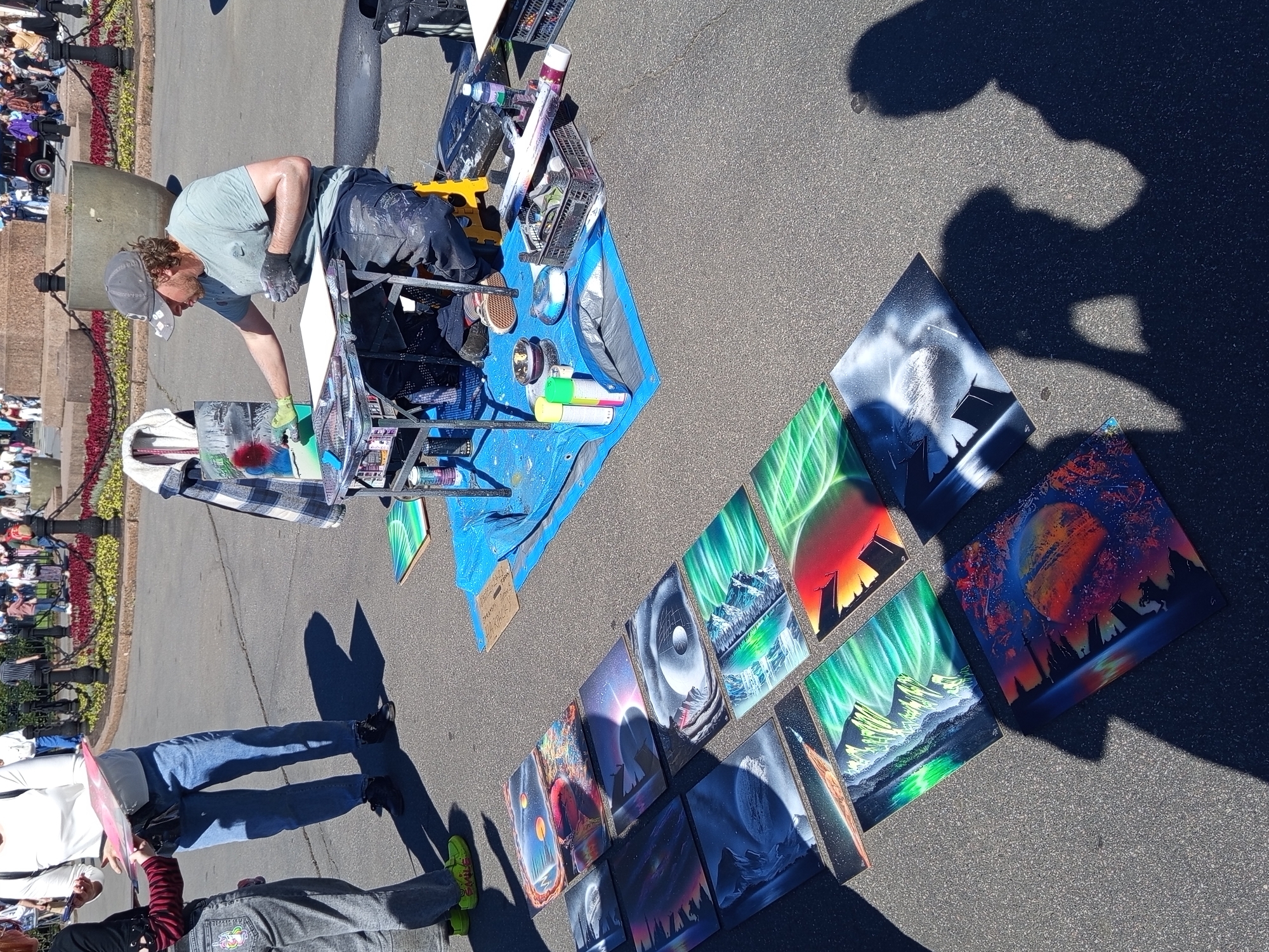 An artist is spray painting a canvas laid on the ground, surrounded by a collection of finished spray-painted artworks displayed in a row on the asphalt. The artist, wearing a cap, t-shirt, and dark pants splattered with paint, is focused on his work, wearing gloves. The setting appears to be an outdoor market or fair, with a crowd of people visible in the background along a walkway with decorative fencing and trees. The artworks themselves are abstract and landscape-themed, featuring vibrant colors and depictions of the aurora borealis, mountains, and celestial bodies. Some of the artworks are leaning against a rolling cart laden with spray paint cans and art supplies. The overall scene suggests a sunny day, with strong shadows cast by the people and artworks, indicating midday. The location context of Saint Petersburg, Russia, is not directly evident from the visual cues in the image. There is no visible text within the frame.