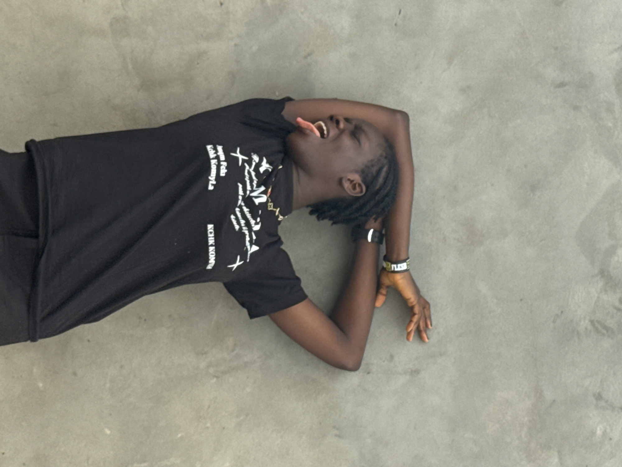 A young adult lies on their back on a flat, gray surface, presumably a floor. The person, seemingly in distress, has their arms crossed above their head, tongue extended out of their mouth. They are wearing a black t-shirt with white print, and two wristbands are visible on their right wrist. The location, Badagry, Nigeria, cannot be determined from the image alone.