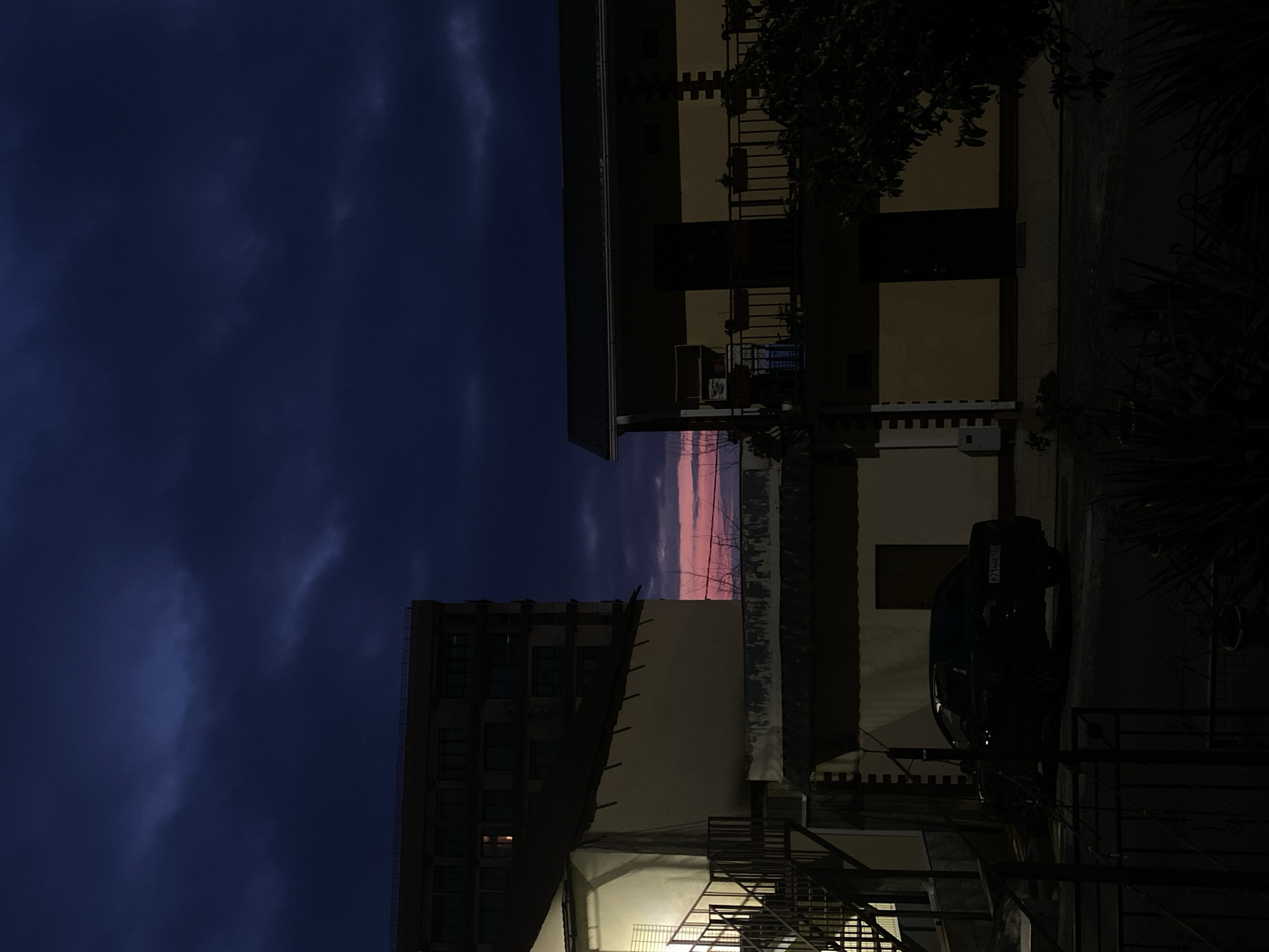 The image depicts a nighttime scene in an urban environment, presumably in Sochi, Russia. A narrow band of pink and red light is visible on the horizon between two buildings, suggesting either sunrise or sunset. The sky is predominantly a deep blue with visible cloud formations.
On the right, a light-colored building with balconies and a visible staircase is partially illuminated. A dark-colored car is parked adjacent to the building, bearing a license plate. The license plate appears to have the alphanumeric code "P.7 LOVE 123". Vegetation is visible near the building. On the left, a tall, multi-story building with lit windows is captured in silhouette. An external staircase is present on the side of the building facing the camera.