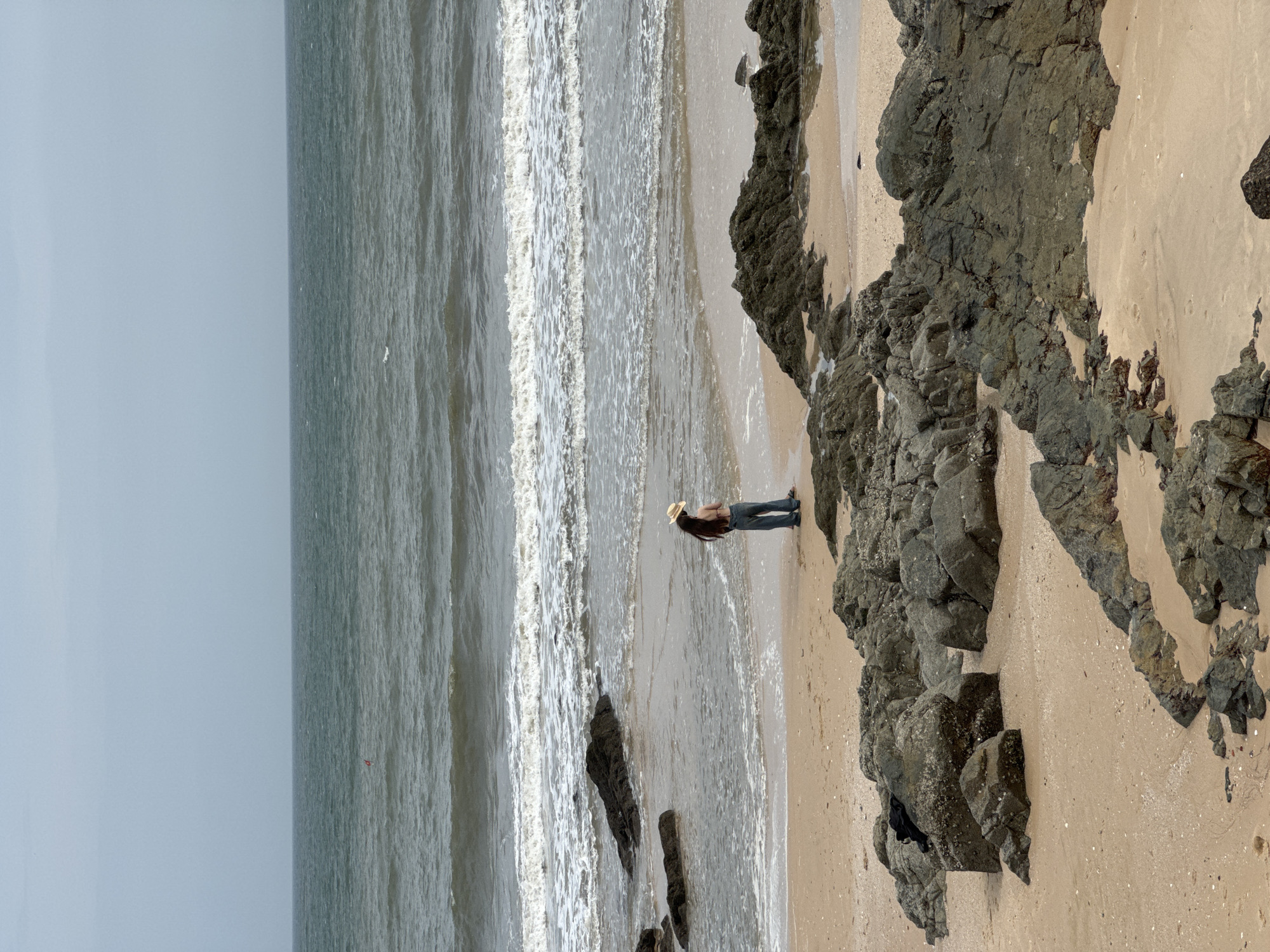 A person stands on a sandy beach in Mũi Né, Vietnam, facing the ocean. The individual wears a straw hat, a light-colored top, and dark pants. They are positioned near the water's edge, with waves breaking on the shore. The beach is dotted with large, dark rocks. In the distance, the sea stretches to the horizon under an overcast sky. A small red buoy is visible in the water. The scene captures a moment of solitude by the sea.