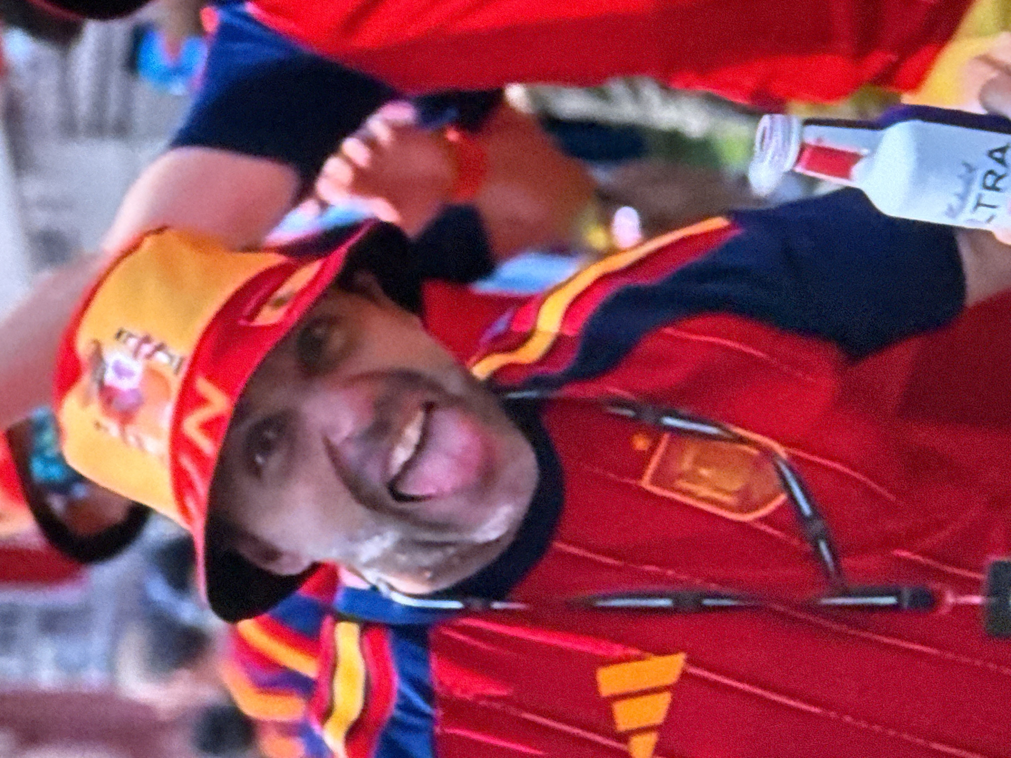 The image shows a close-up of a man wearing a red and yellow bucket hat and a red soccer jersey. He is smiling with his mouth open and his tongue sticking out slightly. The hat has a design on it that includes red and yellow elements and what appears to be text or a logo. The jersey has red and blue accents and a crest or logo on the chest. The man is holding a white bottle with red accents and text that reads "ULTRA" in what appears to be a drinking establishment or sports event. The background is blurred but suggests a crowded outdoor environment with people in similar attire, indicative of a sporting event or celebration. There is no information in the image to suggest the location is Malmyzh, Russia.