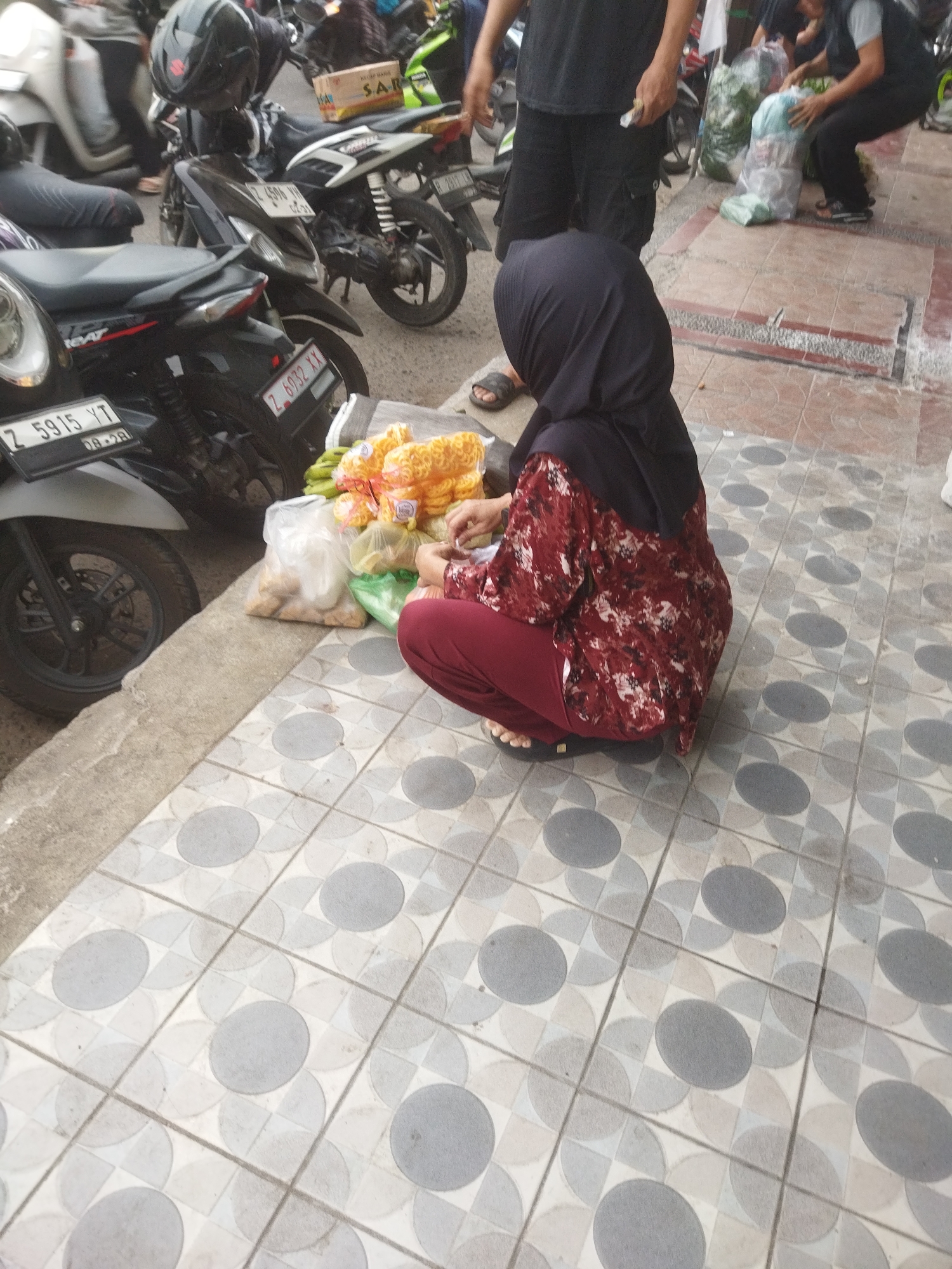 A woman wearing a black hijab and a red and white patterned shirt with maroon pants crouches on a tiled sidewalk. She is sitting next to bags of snacks, including what appears to be puffed corn snacks and bananas. Behind her, several motorcycles are parked. The woman is looking down at the snacks and her hands are near them. The sidewalk is patterned with gray circles and squares. The scene is outdoors, likely on a street in Banjar, Indonesia, given the presence of parked motorcycles and the style of the woman's attire.