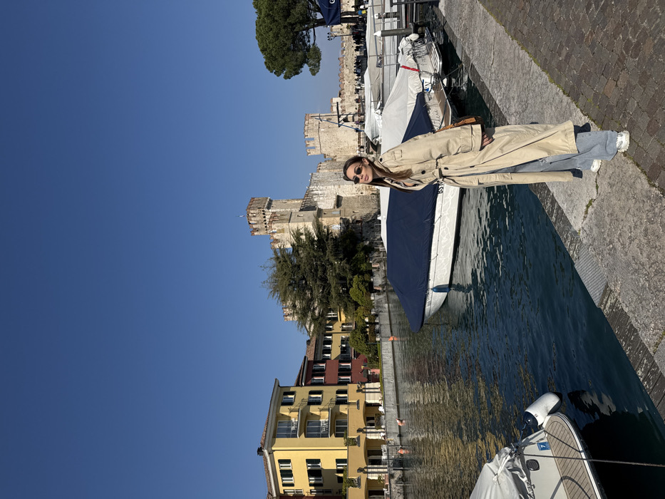 An outdoor scene in Sirmione, Italy, depicts a canal alongside a stone-paved walkway, with the medieval Scaliger Castle (Rocca Scaligera) in the background.
A female individual stands on the right side of the stone walkway, facing slightly left towards the camera. She wears dark sunglasses, a light beige trench coat, light blue wide-leg jeans, and white sneakers. A small brown shoulder bag is visible. Her dark hair is down, and her expression is neutral.
To her left, a narrow canal with calm, blue-green water runs parallel to the walkway. Several boats are moored in the canal; a white boat with a dark blue cover is prominent in the center, and the stern of another white boat with a white cover is visible in the far left foreground. The water reflects the clear blue sky.
Across the canal, a row of multi-story buildings with colorful facades (yellow, red, orange) and balconies line the bank. Behind these buildings, and dominating the central and right background, stands the Scaliger Castle, characterized by its stone walls, crenellated towers, and archways. Green trees are situated around the castle structures and to the far right. Distant figures of people are visible near the castle's entrance. The sky overhead is clear and deep blue.