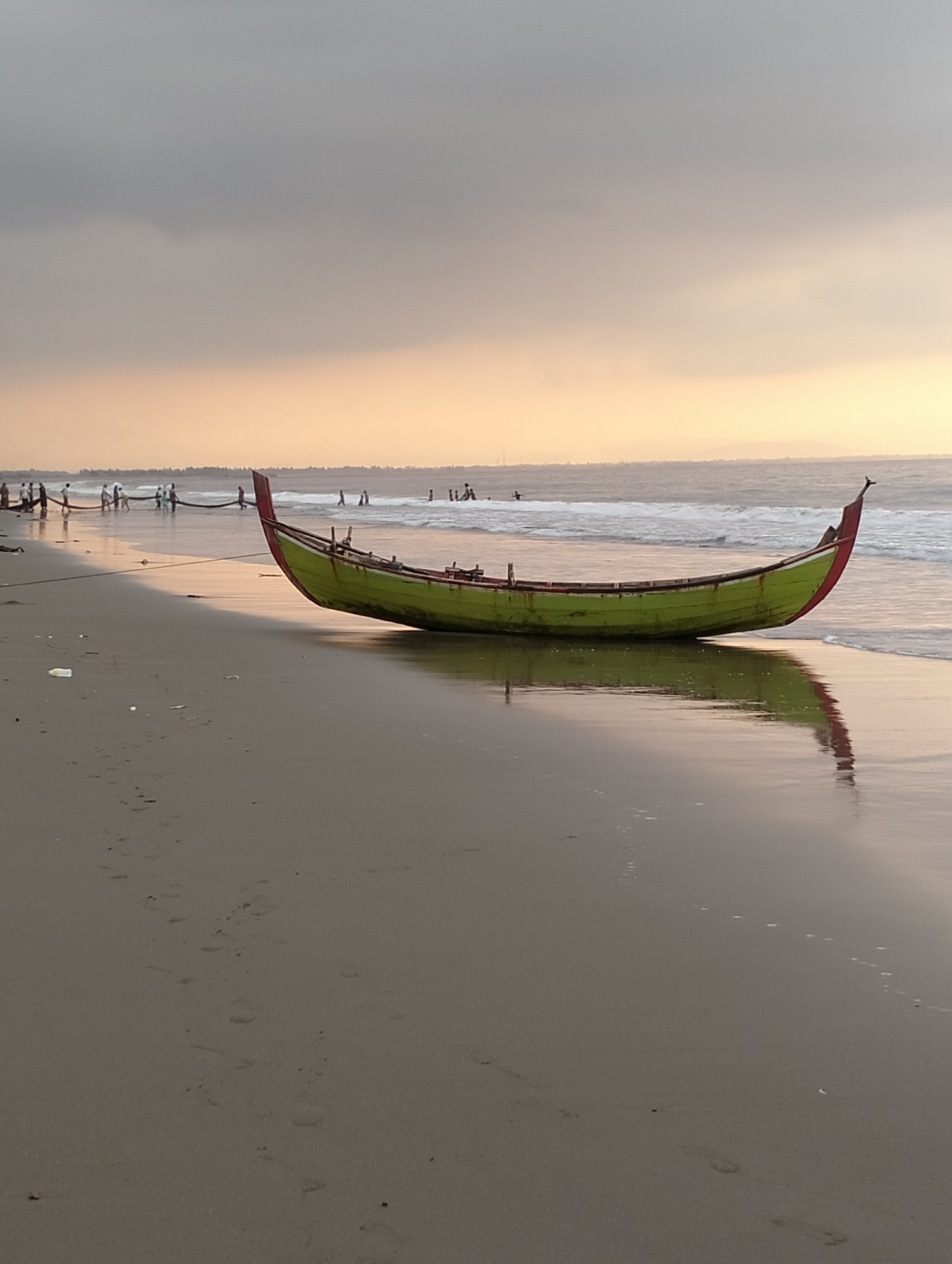 A long, narrow, green and red fishing boat is beached on the wet sand of a coastline. The boat is angled from lower left to upper right. The bow of the boat is to the right and points towards the sea. The stern of the boat is to the left and is closer to the viewer. The boat is resting in the shallow water, its reflection visible on the wet sand. In the background, a group of people, possibly fishermen, are gathered near the water, some of them pulling a fishing net. The sea is calm with small waves. The sky is overcast with clouds, and the light suggests it is either early morning or late afternoon. The beach is sandy with some scattered debris. The scene depicts a fishing activity at the coast of Peuneulet Baroh, Indonesia.