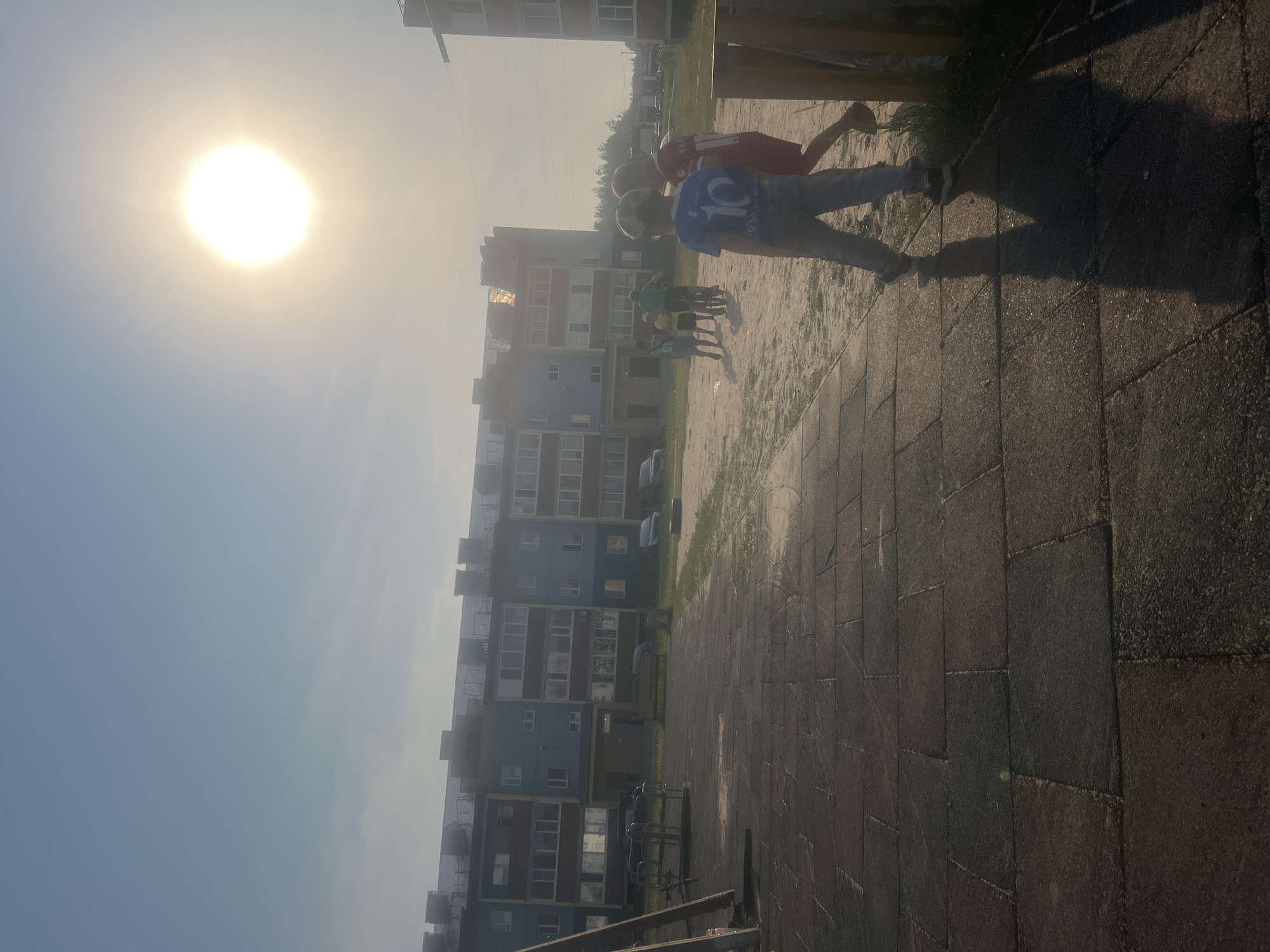 The image depicts an outdoor scene with a bright sun high in the sky, suggesting it is midday. The sun's glare is intense, casting long shadows on the ground. In the foreground, a paved area with large, dark rectangular stones dominates the lower right portion of the frame. To the left, a sandy area is visible, with a line of trees and apartment buildings in the background. The buildings are multi-story structures with balconies and numerous windows, painted in shades of blue and red.
Several people are present in the image, though their faces are not clearly visible due to the angle and lighting. Two individuals are in the mid-ground, with one wearing a blue t-shirt with a white number "10" on the back, and the other in a red shirt with a white number "11." They appear to be engaged in some form of activity, possibly playing or exercising. Further in the background, a group of smaller figures, likely children, are standing on the sand. The overall atmosphere seems casual and perhaps playful, given the presence of children and the bright daylight. The time of day is clearly daytime, and the weather appears to be sunny and clear, though a slight haze might be present due to the sun's intensity. No explicit text is clearly legible.