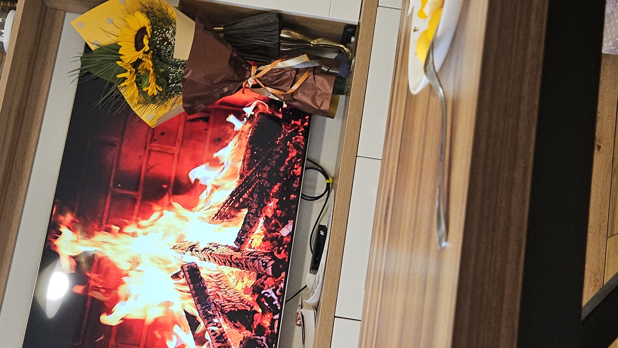 The image shows a bouquet of sunflowers and wrapped objects placed on top of a television screen displaying a fireplace image. The screen is surrounded by a white frame. To the right, there is a wood panel and a thin, silver object. The location is unknown, and there is no indication of being in Plovdiv, Bulgaria. There are no people in the image.