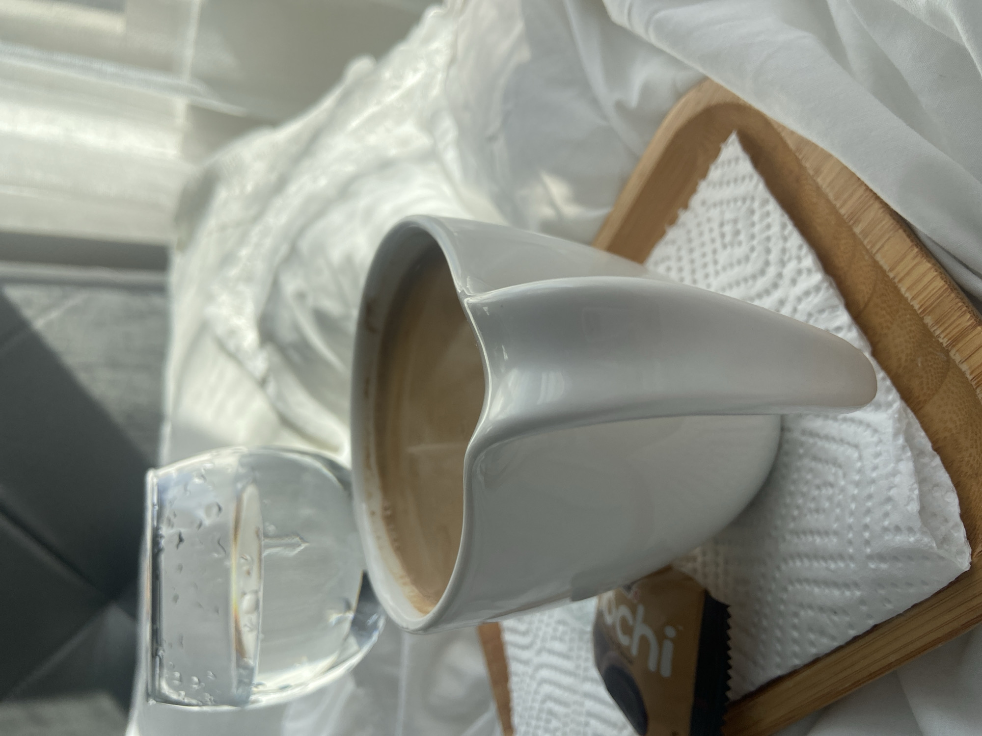 The image depicts a coffee cup on a small wooden tray, next to a glass of water. The tray rests on a white, wrinkled fabric, likely bedding. A small, wrapped snack is also on the tray, partially visible. The scene appears to be indoors, possibly in a hotel or apartment. There is no direct indication of the location being Budva, Montenegro, other than potentially the style of the setting. No people are visible.