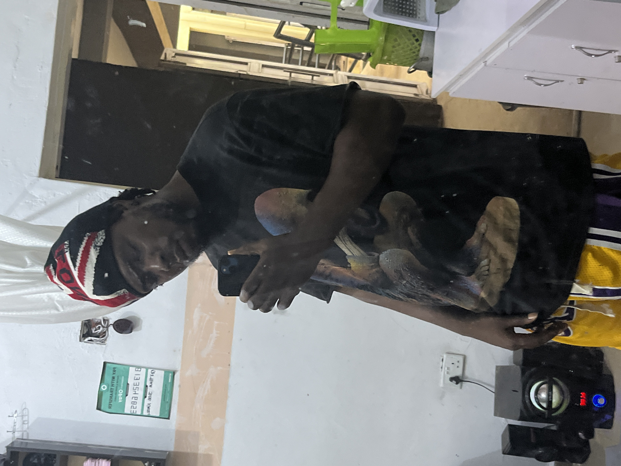A young Black man stands in a room, taking a mirror selfie. He is wearing a black t-shirt with a graphic design on it, a beanie with a red, white, and black pattern, and yellow basketball shorts. He is holding a black smartphone in his right hand. In the background, a white wall is visible, with a green sign that has text in both English and what appears to be an African script. An electrical outlet and a round, dark object are also on the wall. To the left of the man, there is a black speaker system. To the right, there is a white cabinet with silver handles, and a green plastic basket. The room appears to be a domestic space, possibly a bedroom or living area, within a residential building in Jos, Nigeria. The lighting is artificial and somewhat dim. The man's expression is neutral, and he is looking at his phone. The overall impression is of a casual, everyday moment captured in a personal space.