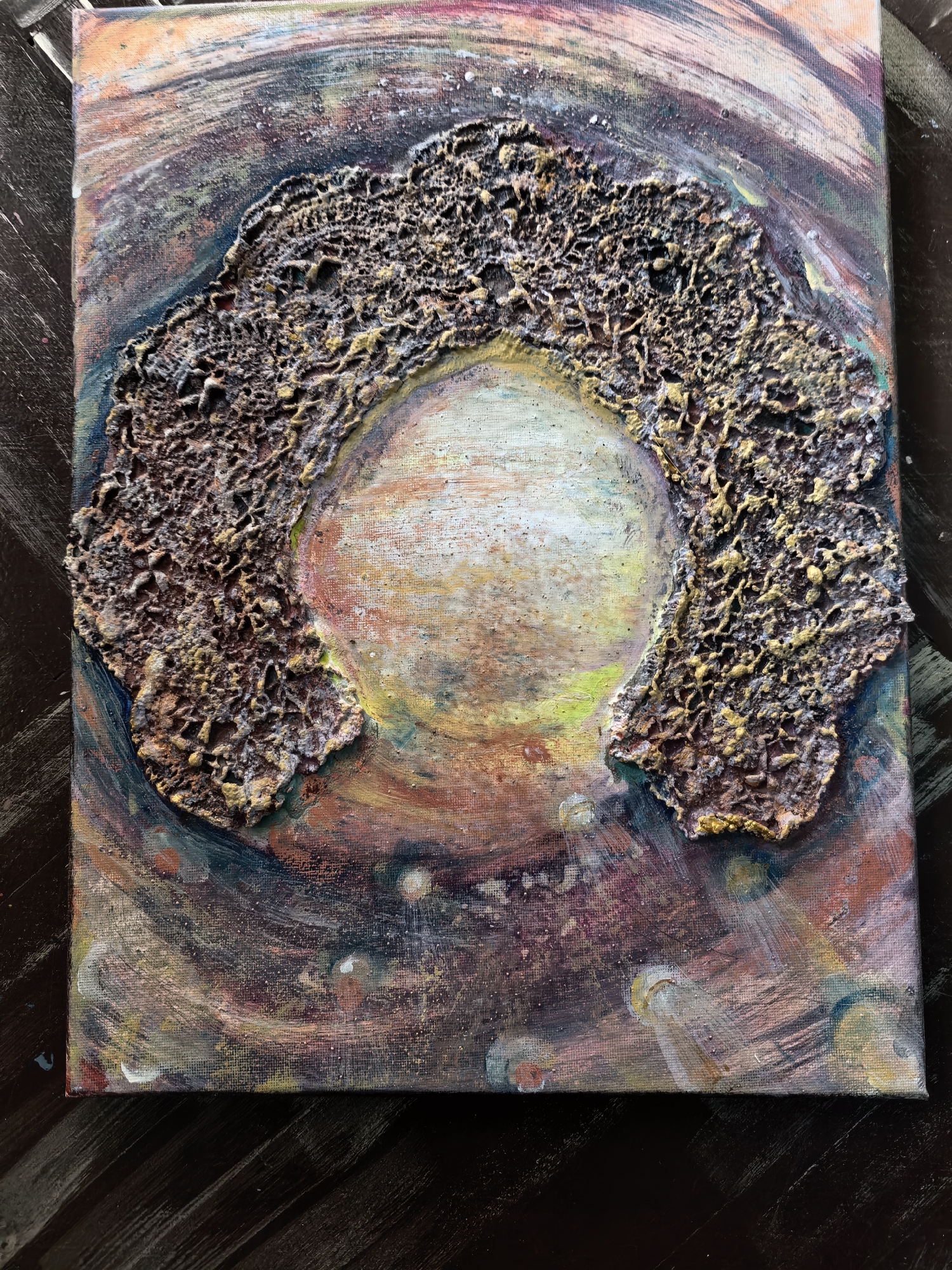 The image is a close-up of an abstract painting on canvas. The central motif is a roughly circular, light-colored area surrounded by a textured, brown-and-gold, somewhat irregular structure that suggests a partial frame or border. The background features swirling patterns and colors, including shades of brown, purple, orange, and blue. The canvas rests on a dark wooden surface. The location and city are unknown.
