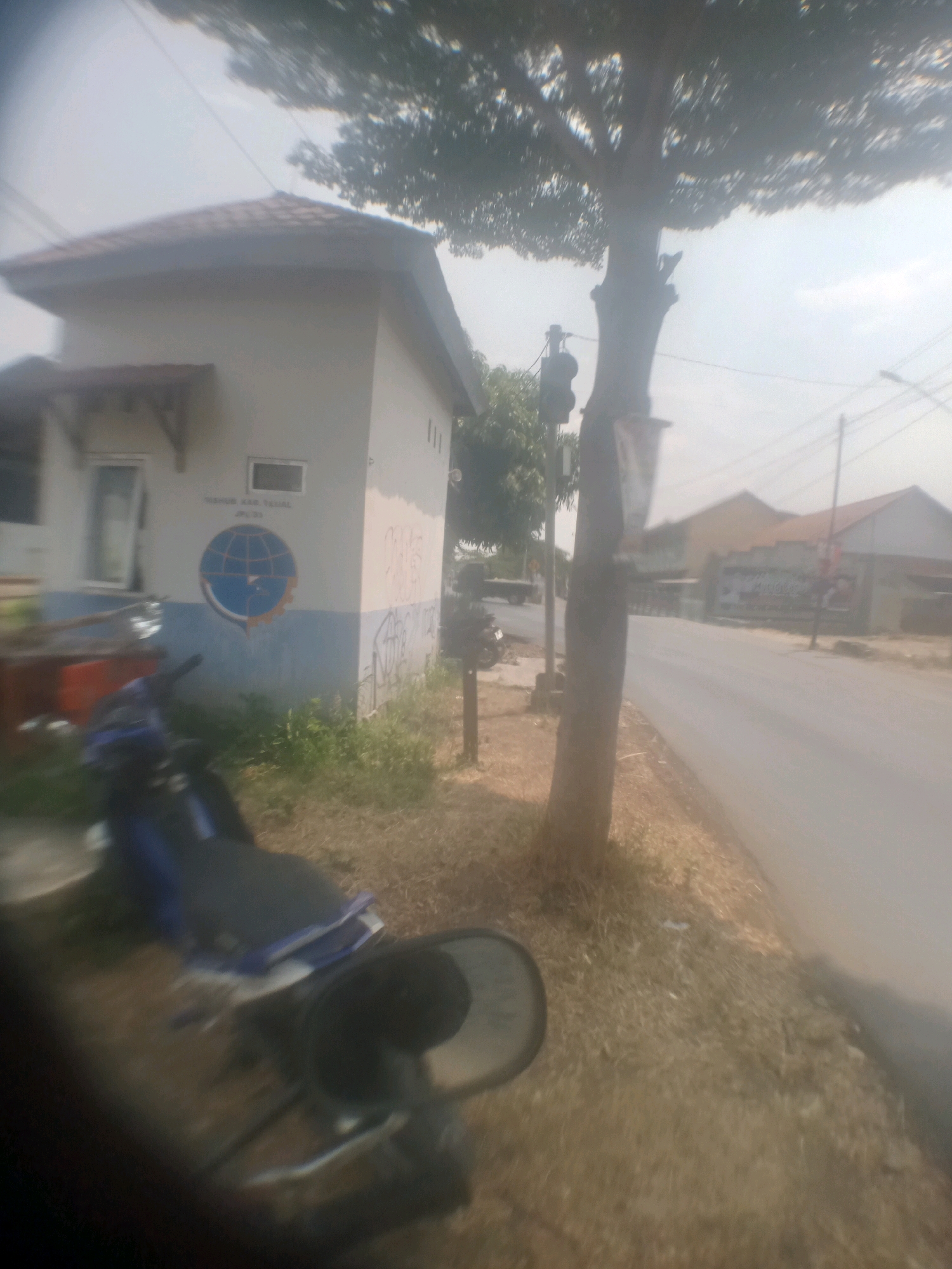 The image depicts a street scene in Lebakgowah, Indonesia. In the foreground, a blue and black motorcycle is parked on a dry, grassy shoulder next to a paved road. Behind the motorcycle, to the left, is a small, light-colored building with a brown tiled roof and a blue and white mural of a globe on its side. The building has at least one window and a small awning. A tall tree with a thick trunk stands beside the building and the road, its branches extending towards the top of the frame. A traffic signal is visible on a pole near the tree. The road stretches into the background, with more buildings visible on the right side, one with a prominent sign. The sky is bright and hazy, suggesting a sunny day. Power lines are visible crisscrossing the sky. The overall scene suggests a quiet, possibly semi-rural or suburban area.