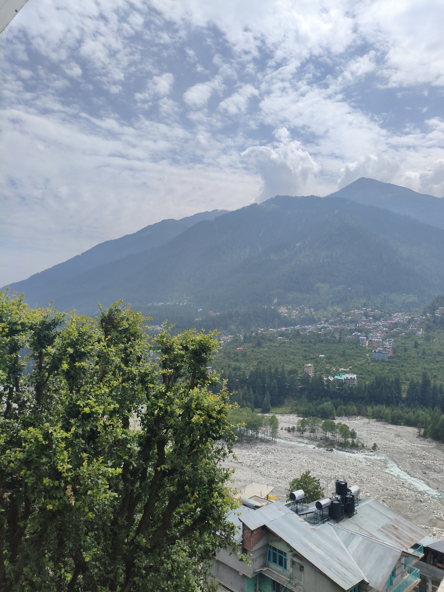 This is a photograph taken in the city of Vashisht, India, showing a scenic view of mountains, a valley, and a river. In the foreground, a large, leafy green tree occupies the left side of the frame, with its branches and foliage obscuring parts of the scene. Below the tree, the roof of a building is visible, constructed with a corrugated metal roof and several water tanks.
Beyond the foreground, the landscape opens up to reveal a wide, rocky riverbed with a narrow, turquoise stream flowing through it. The valley floor is covered in lush green vegetation, with a dense forest of pine trees. Scattered within the forest and on the slopes are buildings, indicating a settlement or village.
In the background, imposing mountains rise, their peaks shrouded in a light haze. The sky is partly cloudy, with formations of white clouds against a pale blue backdrop. The overall impression is one of a natural, mountainous landscape with signs of human habitation in the valley.