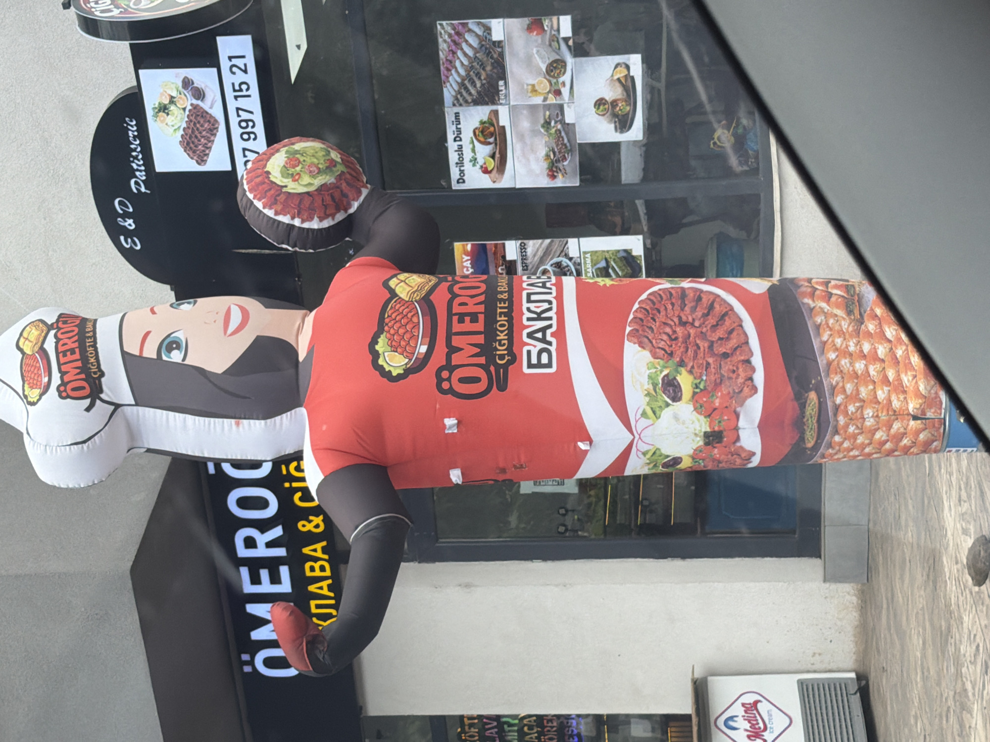 In Aytos, Bulgaria, an inflatable mascot wearing a chef's hat and a red shirt is positioned in front of a restaurant with a glass storefront. The mascot is holding a circular object, possibly food. The restaurant sign above the mascot reads "ÖMEROĞLU ÇIĞKÖFTE & BAKLA". Another sign on the right side of the storefront displays various food items, including what appears to be döner and baklava. A smaller sign to the left of the main entrance indicates "Medina ice cream". The ground in front of the establishment is paved, and a single dark-colored rock is visible on the ground. The overall scene appears to be an outdoor commercial area.
