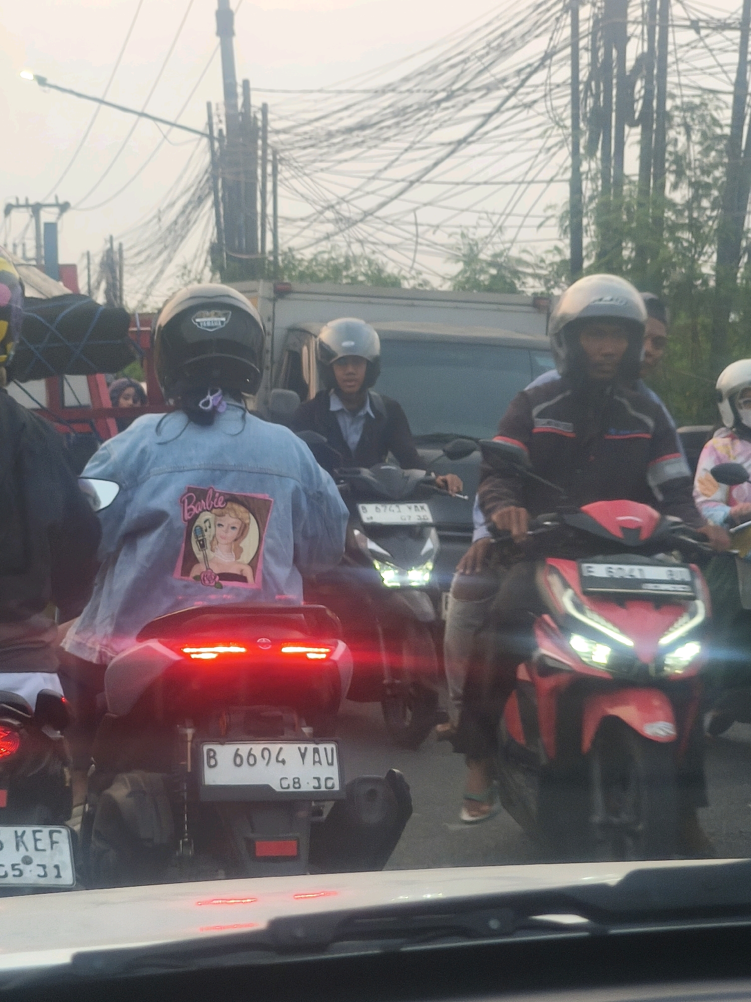The image depicts a traffic jam on a hazy day in Bekasi, Indonesia. Several motorcycles are visible, carrying passengers wearing helmets and protective gear. One rider in the foreground is wearing a denim jacket with a large Barbie graphic on the back. Another rider, to the right, is on a red motorcycle. The background shows a dense network of electrical wires overhead, indicating an urban environment. The overall scene is one of congested urban traffic.