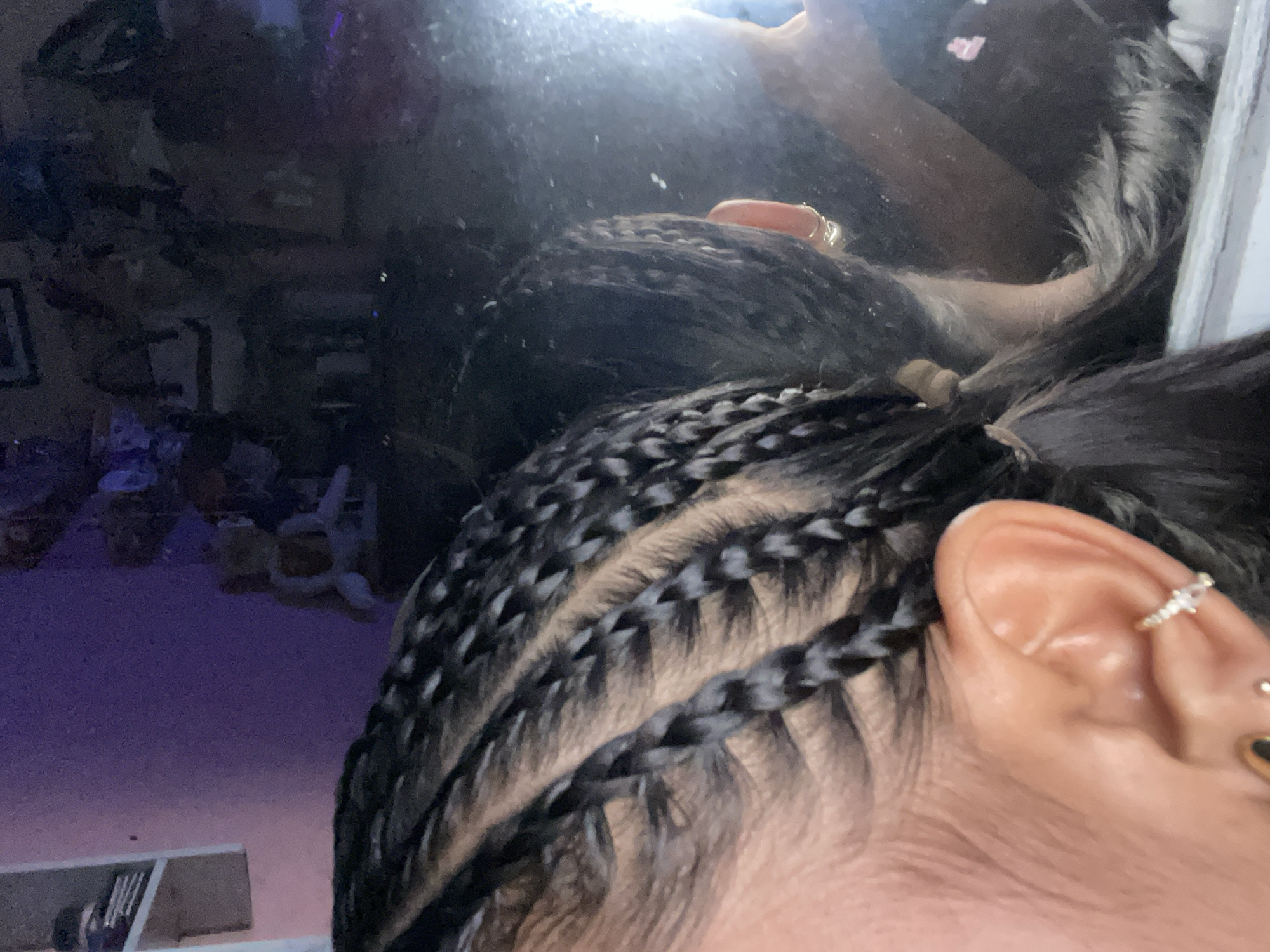 Here is a close-up shot taken in an indoor environment. The focal point is on a person's ear and a portion of their head with intricate braided hair. The individual has multiple ear piercings with jewelry visible in each. The hair is styled in close braids that run along the scalp, with the ends gathered. The background includes a reflection of a room with what appears to be furniture and decorative items. The lighting in the image is a mix of natural and artificial, creating some glare in the reflection.