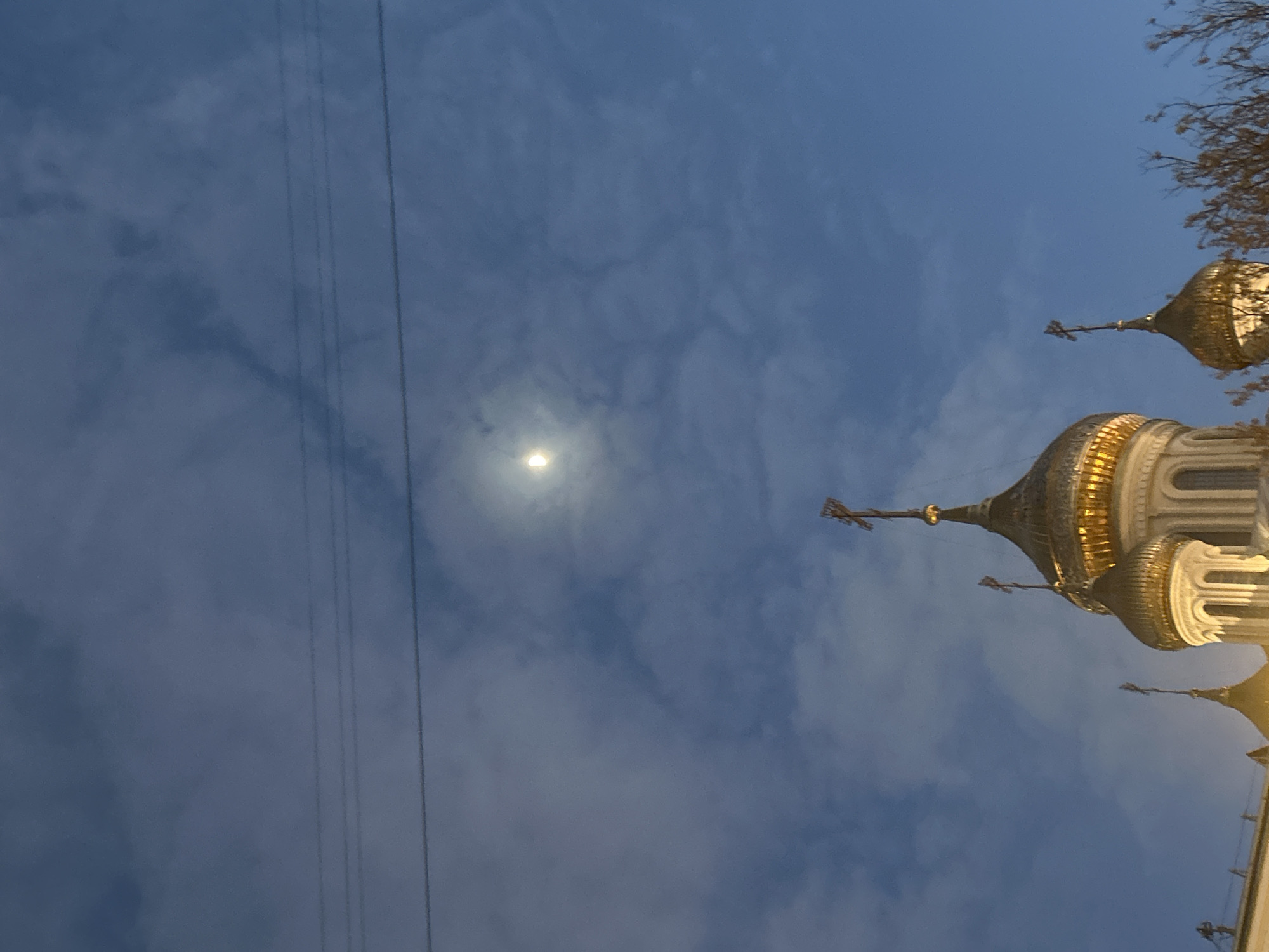 The image displays a dramatic architectural view against a cloudy, deep blue sky, likely at dusk or night. The main subjects are the ornate, golden onion domes of an Orthodox church, positioned on the right side of the frame. Several domes are visible, topped with distinctive Orthodox crosses, and their golden surfaces reflect light. Below the domes, parts of the church's white, classical architectural structure are also discernible.
On the left side of the image, a bright, circular celestial body, appearing as the moon, is visible through scattered clouds. Thin, dark lines, likely power lines or tram wires, cut across the left and central sky. In the upper right corner, bare tree branches are visible, suggesting an autumn or winter season. The overall scene is static, focusing on the grandeur of the architecture under a atmospheric sky. The style of the church architecture is characteristic of Moscow, Russia. There are no people or visible text in the image.