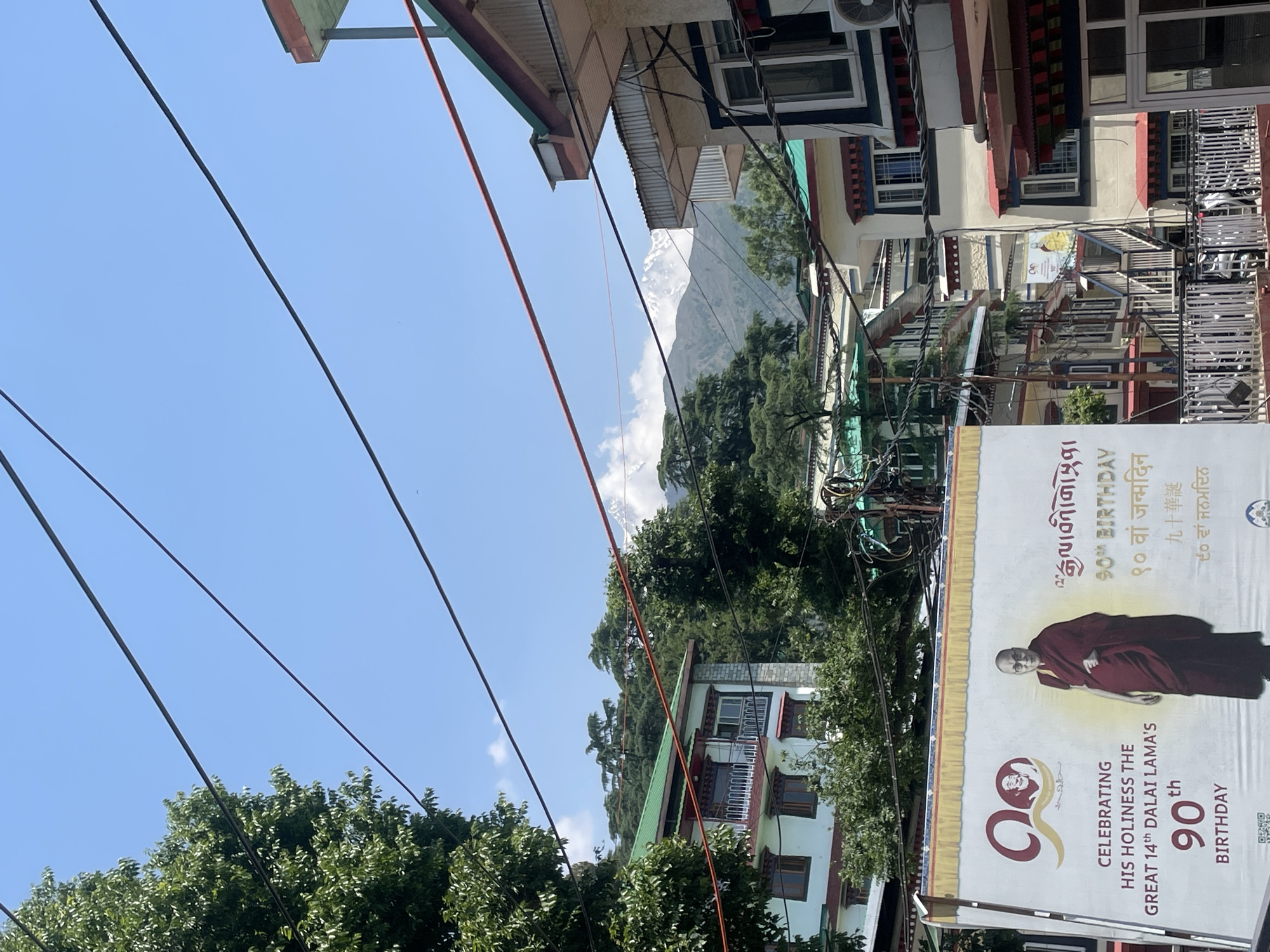 The image presents a view in Dharamshala, India, characterized by a mix of natural and urban elements. In the distance, a snow-capped mountain peak is visible, partially obscured by clouds. Overhead, numerous power lines intersect the sky. Buildings with multi-tiered roofs and multiple windows are constructed on the sides. On the right, a large banner announces the 90th birthday celebration of the 14th Dalai Lama, featuring his image and text in multiple languages. Lush green trees are scattered throughout the scene. The photo appears to have been taken from an angle, giving the impression of a tilted horizon.
