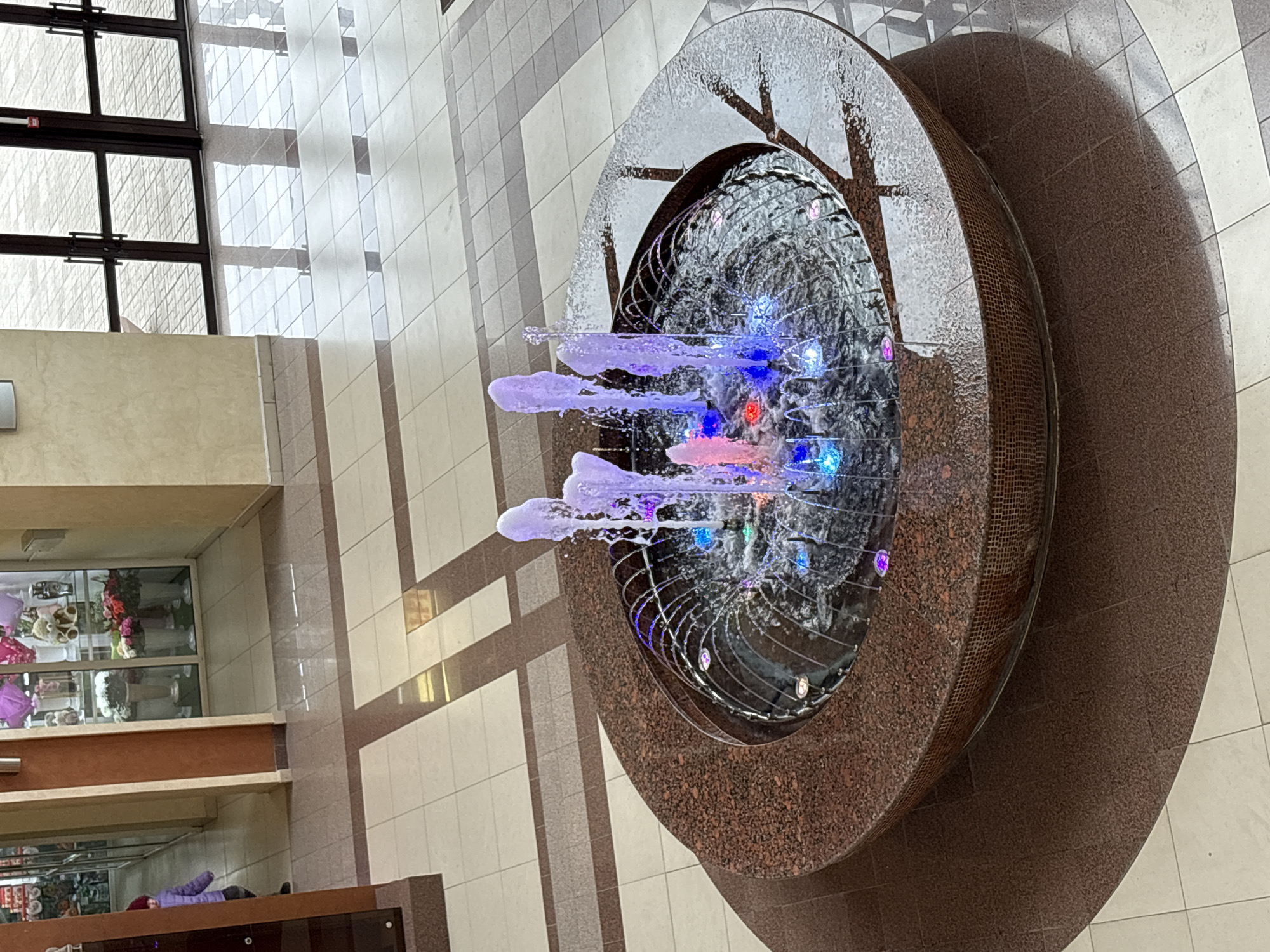 An indoor fountain featuring illuminated water jets is positioned in an open area with tiled flooring and vertical beige and brown patterned walls. The fountain consists of a round, tiered base made of a dark speckled material. The fountain's inner basin contains water jets illuminated in multiple colors including blue, pink, and purple. The floor tiles alternate in shades of beige and gray, creating a grid pattern. The walls feature vertical stripes of beige and brown. A windowed storefront is visible in the background, displaying floral arrangements and plush toys. Above the storefront, a set of dark-framed windows is seen. The location is in an interior space in the city of Unknown, Unknown.