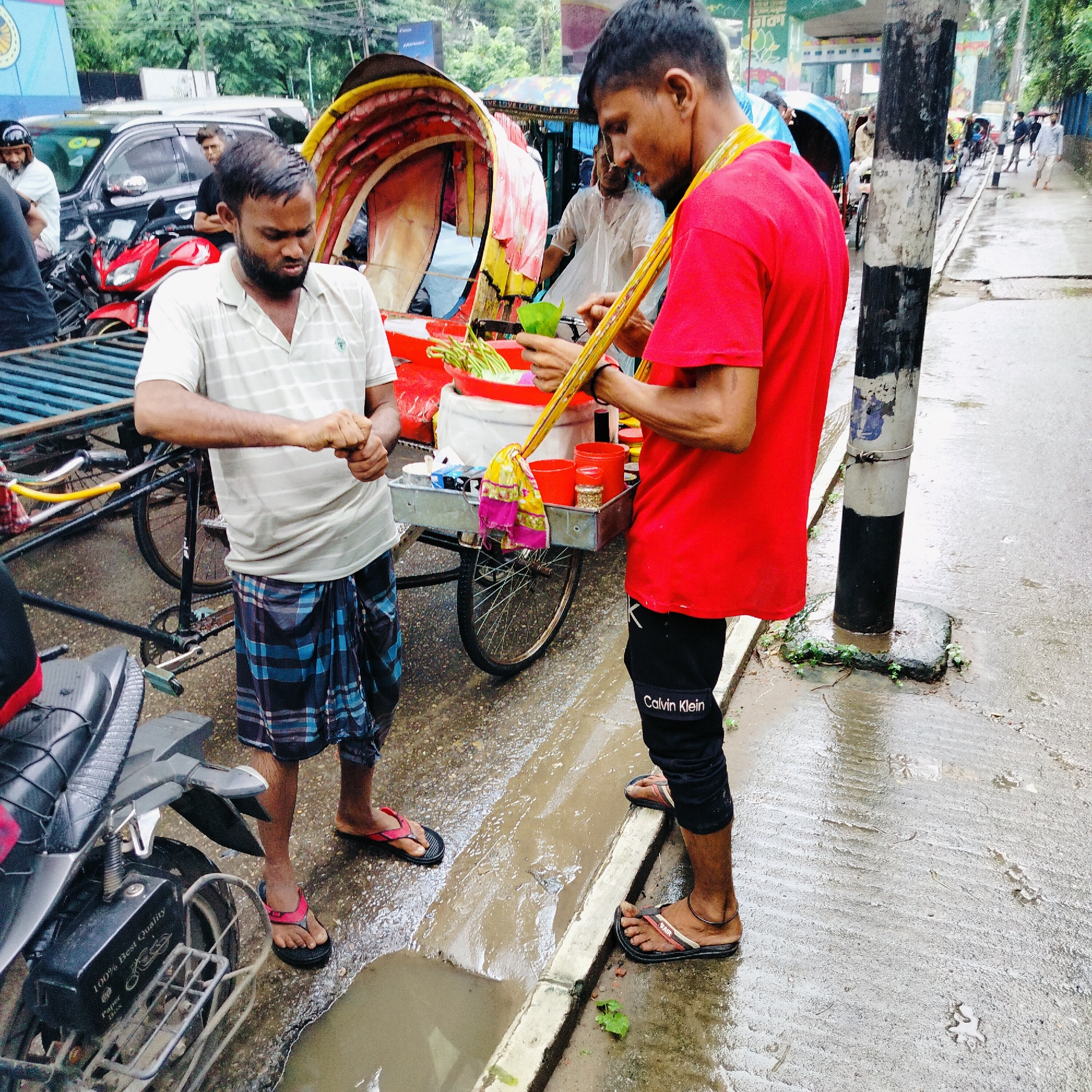 Two men are standing next to a roadside cart in Dhaka, Bangladesh. The cart appears to be selling betel leaf preparations, as indicated by the green leaves and other items in red containers. One man, wearing a red t-shirt and black pants with "Calvin Klein" printed on them, is holding a yellow stick adorned with a colorful cloth and a green betel leaf. The other man, dressed in a striped polo shirt and a blue checkered lungi, is facing away from the camera and appears to be handling items from the cart. The ground is wet, suggesting it has rained recently, and there are puddles of water visible. In the background, a red motorcycle and a rickshaw are parked, along with other vehicles and people, indicating a busy urban environment. The overall scene depicts a typical street-side vendor operation in Dhaka.