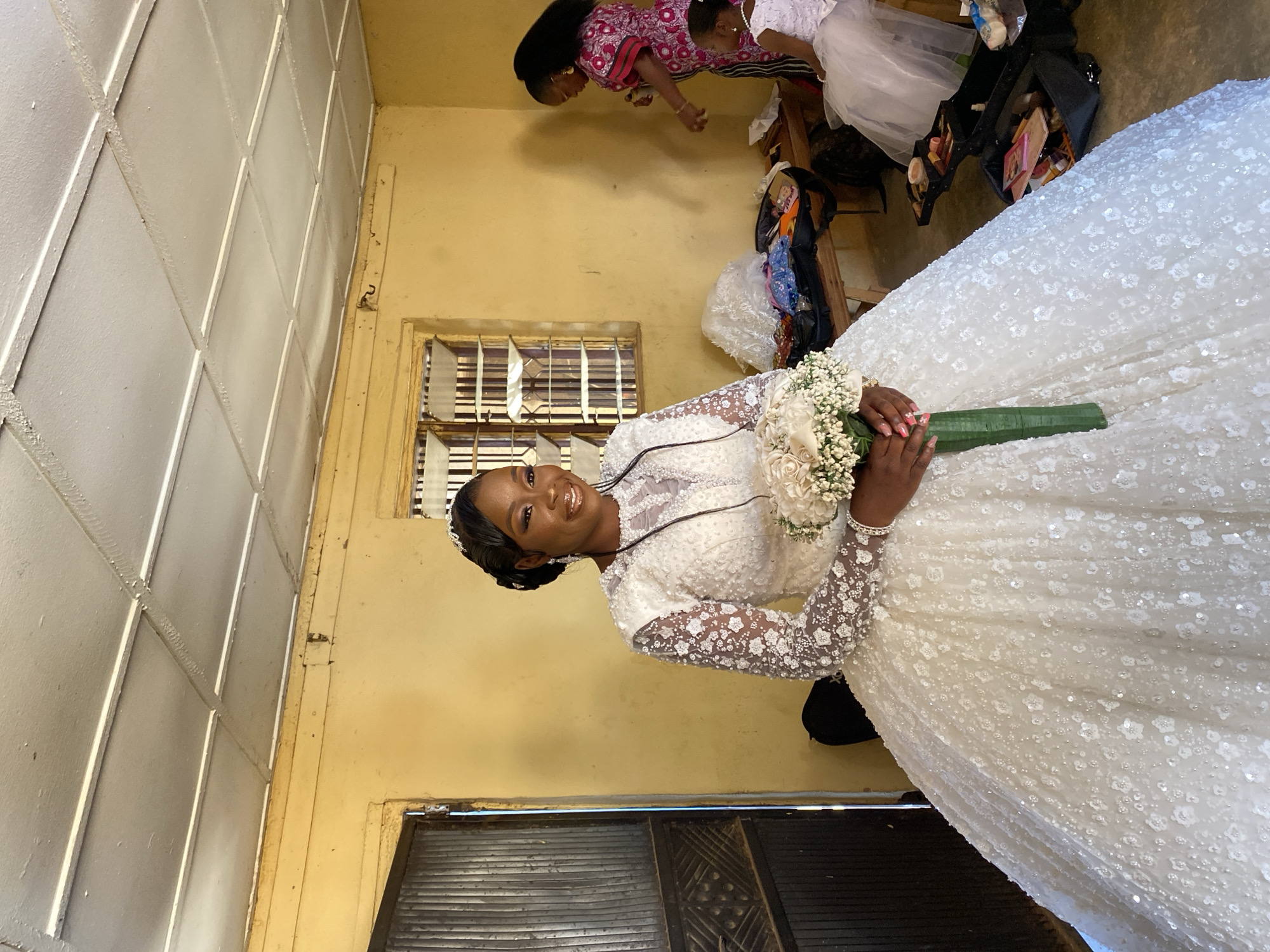 In Vom, Nigeria, a woman in a white, long-sleeved, embellished wedding dress stands indoors, holding a bouquet of white flowers. The room has yellow walls and a window with metal bars. A partial view of a doorway is seen. In the background, partially obscured, are two other women and various items like a garment bag and makeup kit.