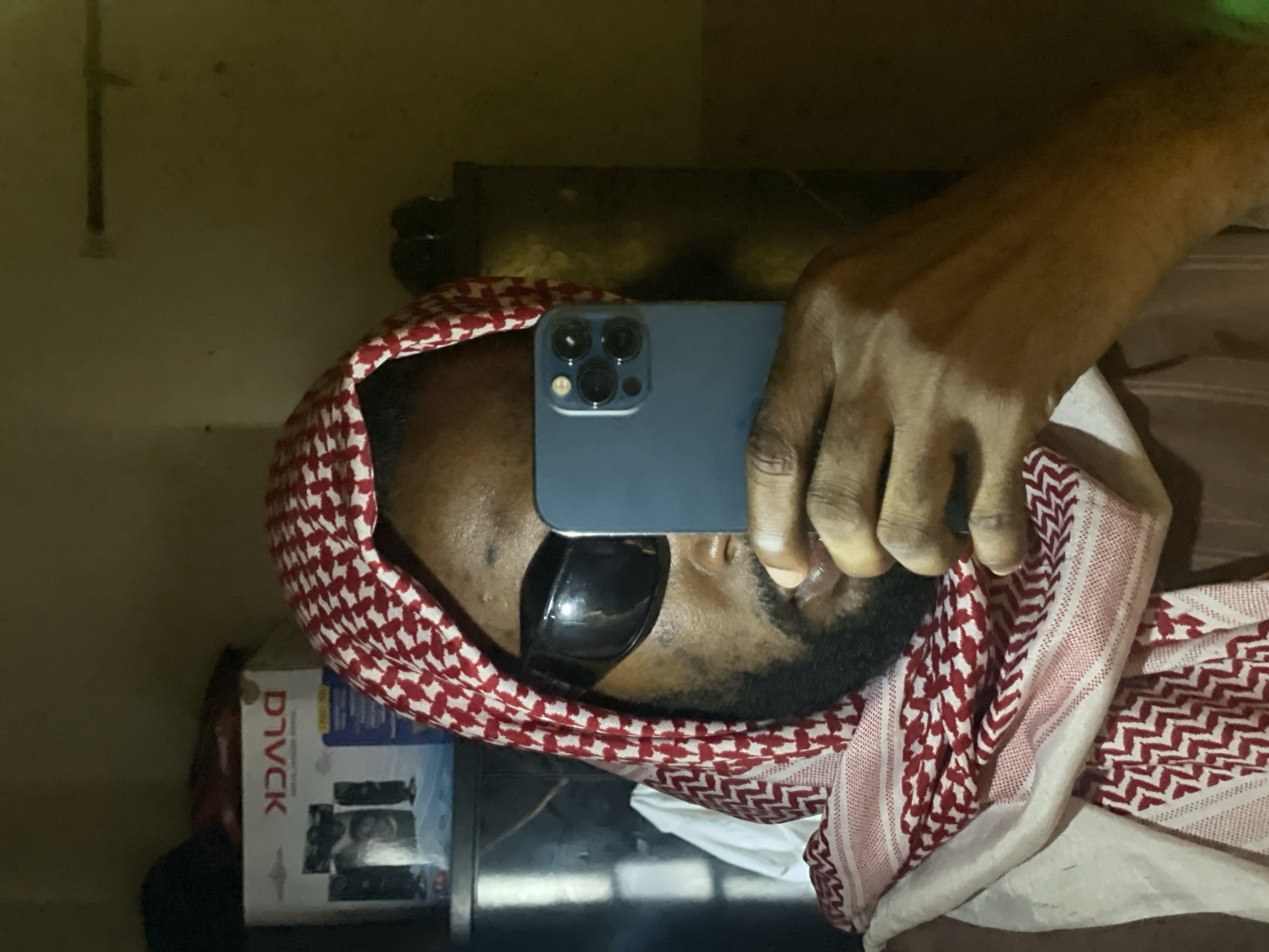 A man wearing a red and white checkered keffiyeh and dark sunglasses is holding a blue smartphone in front of his face, seemingly taking a selfie. A white box labeled "DVLCK" with images of audio equipment is visible near the left side of the image. The background is dark and indistinct. There is no evidence suggesting the location is Lokoja, Nigeria.