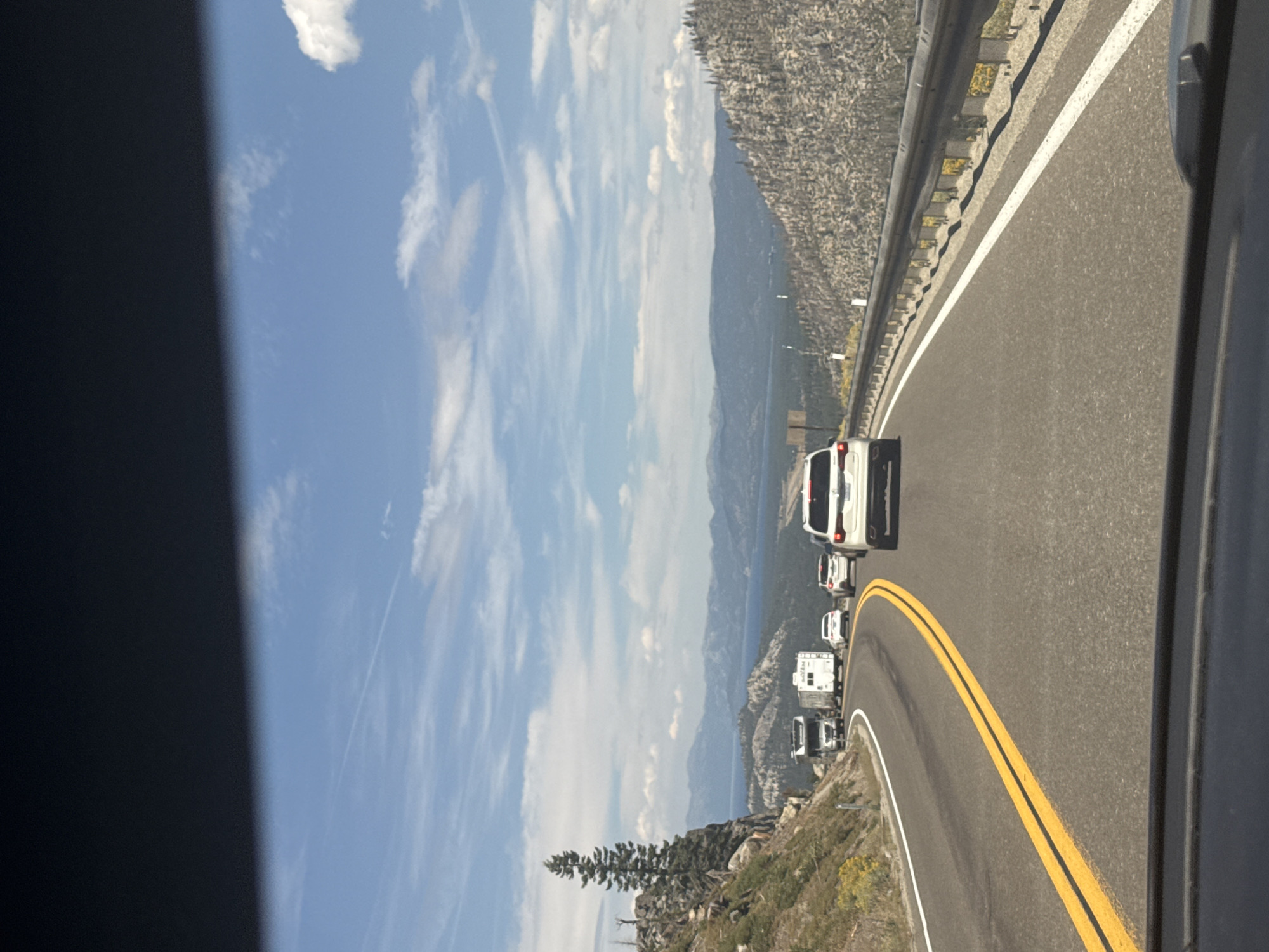 The image shows a view from a car driving on a highway in South Lake Tahoe, United States. The highway curves to the left, with a steep, rocky, and vegetated mountainside on the left and a metal guardrail on the right. In the distance, a large lake is visible, surrounded by mountains under a partly cloudy blue sky. Several vehicles are on the highway, including a large truck towing a trailer, an RV, and several passenger cars. The overall scene depicts a scenic mountain highway route with traffic.