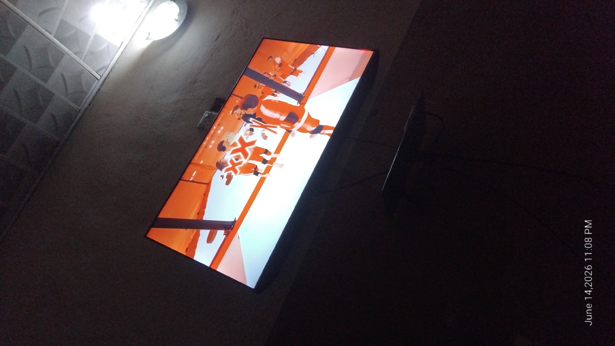 The image shows a television screen displaying a video game. The game features cartoonish figures wearing orange and white outfits, running on a white platform with an orange background. The television is mounted on a wall in a dimly lit room. The ceiling, with its textured tiles and light fixture, is visible in the upper portion of the image. A narrow shelf or mount is visible below the television. The timestamp "June 14, 2026 11:08 PM" is displayed in the bottom left corner of the image. The overall scene suggests a casual, indoor setting, possibly a living room, at night.