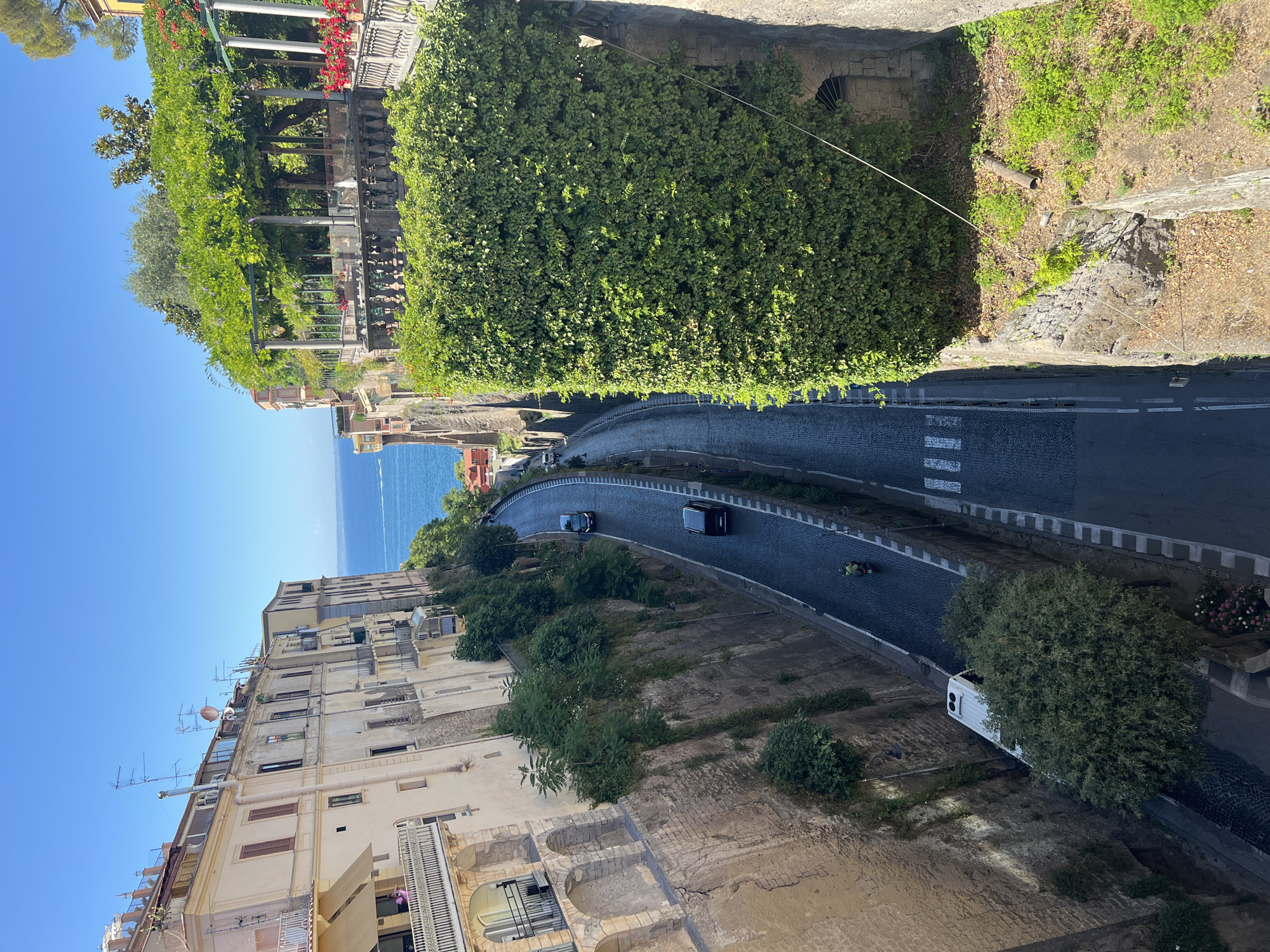 An eye-level medium shot captures a street winding down a coastal cliff in Sorrento, Italy, under a clear blue sky. On the left, a multi-story building with balconies and arched windows stands tall, its facade a muted yellow. Vegetation spills over the edges of the cliff, interspersed with retaining walls. A white bus is parked on the left side of the street near the bottom of the frame.
The street itself is paved with dark cobblestones and is marked with white lines. Two black cars are driving down the street, one behind the other, and a person on a green scooter is also visible further down. On the right side of the street, a dense wall of green ivy covers a cliff face, leading up to a shaded terrace area with pergolas and more greenery, suggesting a restaurant or garden. In the distance, the blue sea stretches out to the horizon. The overall impression is one of a vibrant coastal town nestled on a dramatic cliffside.
