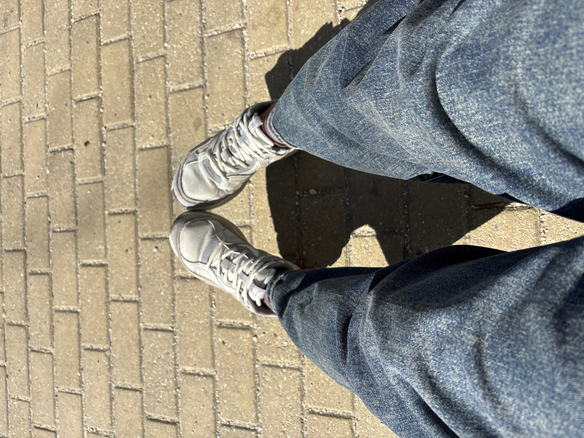A first-person perspective shot shows a person's legs and feet wearing blue denim jeans and gray athletic shoes on a light brown brick walkway. The shoes have white laces and the brand name "FILA" is visible on the side of the right shoe. The brick pathway is illuminated by sunlight, casting shadows from the person's legs and shoes. The environment appears to be an outdoor urban setting, possibly a park or a pedestrian area in Zapadny, Russia. The image is a still shot, with no discernable action or interaction taking place.