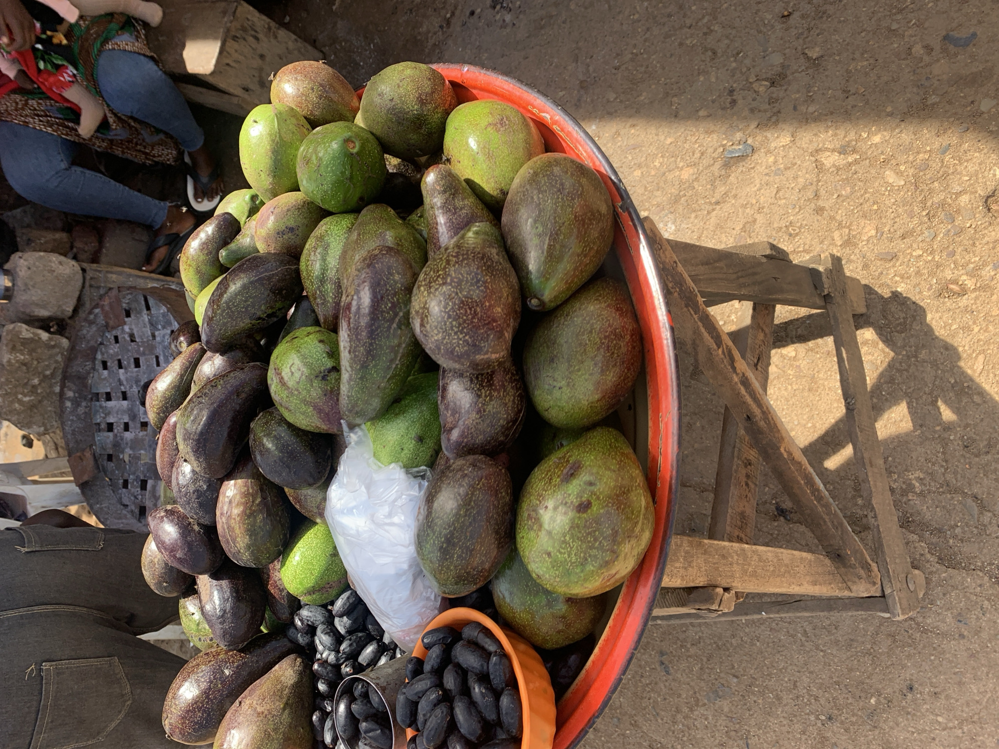The image depicts a market stall in Rafin Par, Nigeria. In the foreground, a large, round, red-rimmed tray is positioned on a rustic wooden stand. The tray holds a substantial quantity of avocados, varying in color from green to purplish-brown, indicating different stages of ripeness. To the left of the avocados, a smaller orange bowl contains numerous small, oval-shaped black fruits, likely African plums (Dacryodes edulis), with additional black fruits scattered directly onto the tray. A crumpled clear plastic bag rests among the produce.
In the background on the left, a person is partially visible from the waist down, wearing dark denim trousers with visible back pockets. On the right, another individual is seated, wearing blue jeans and a patterned fabric draped over their lap, holding a small child whose bare feet are visible. The ground beneath the stand is an uneven dirt or gravel surface, illuminated by strong sunlight casting distinct shadows from the stand and surrounding elements. Stone or concrete blocks are visible in the far left background.