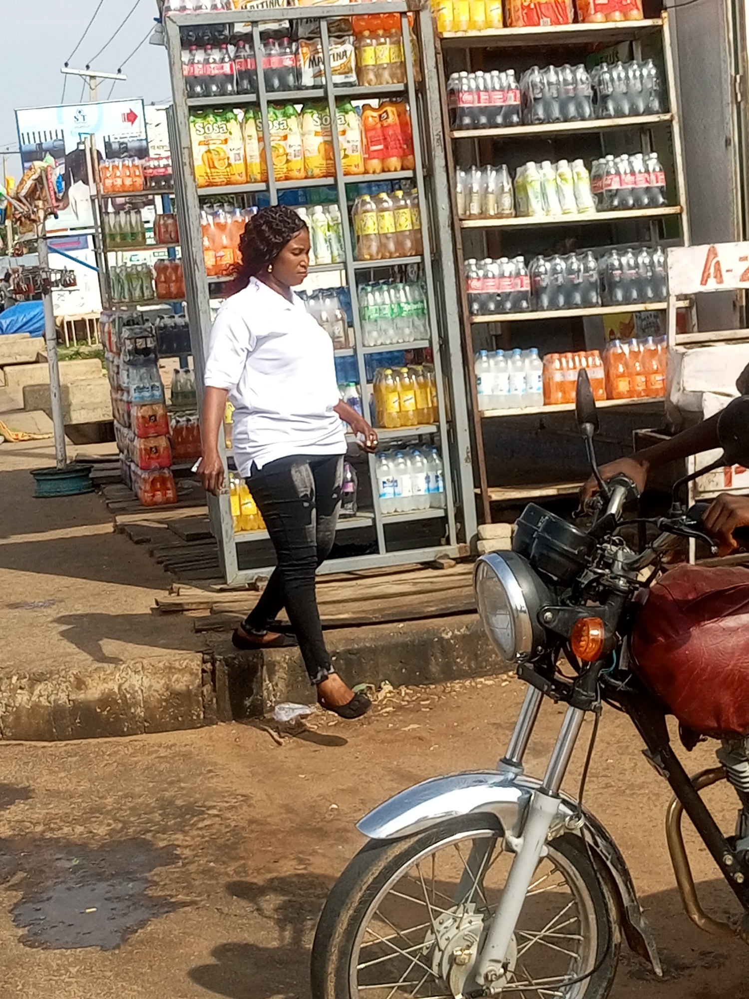 The image depicts an outdoor scene in Gombe, Nigeria, likely near a roadside market. A woman with dark, braided hair is walking past a large display rack filled with bottled beverages. She is wearing a white t-shirt and black jeans. The display rack is stocked with various brands of sodas and juices, some in multipacks. To the right of the woman, the front section of a red motorcycle is visible, suggesting the presence of a rider or that the motorcycle is parked nearby. The ground is a mixture of dirt and some pavement, with shadows cast by the display rack and other unseen structures. The overall environment suggests a busy commercial area.