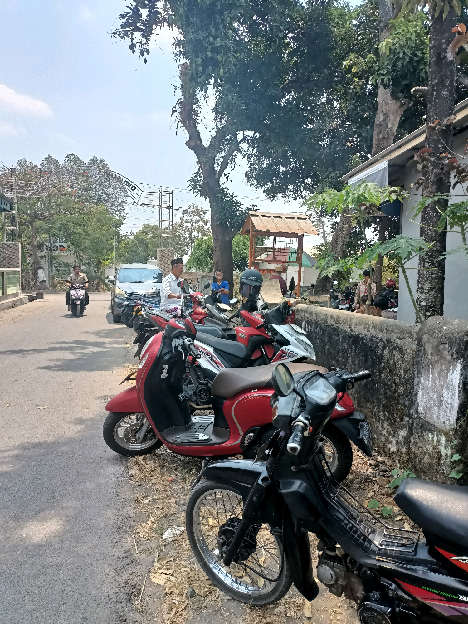 This is a photograph taken on a street in Badas, Indonesia. A row of parked motorcycles lines the side of the asphalt road. In the foreground, a black motorcycle is parked, with its front wheel and handlebars visible. Behind it, a red scooter and other motorcycles are also parked. A low, weathered stone wall runs along the right side of the image, separating the road from a building with a white roof and a dark-tiled section. Several people are visible. A man in a white shirt and peci hat is seated on a motorcycle, looking to his right. Another man in a blue shirt stands near the wall, also looking to his right. Further down the road, a silver car is parked, and a man is riding a motorcycle. Trees and vegetation are present, with a large tree casting shadows on the road. A metal archway with text is visible in the background, above the trees. The sky is bright and clear.