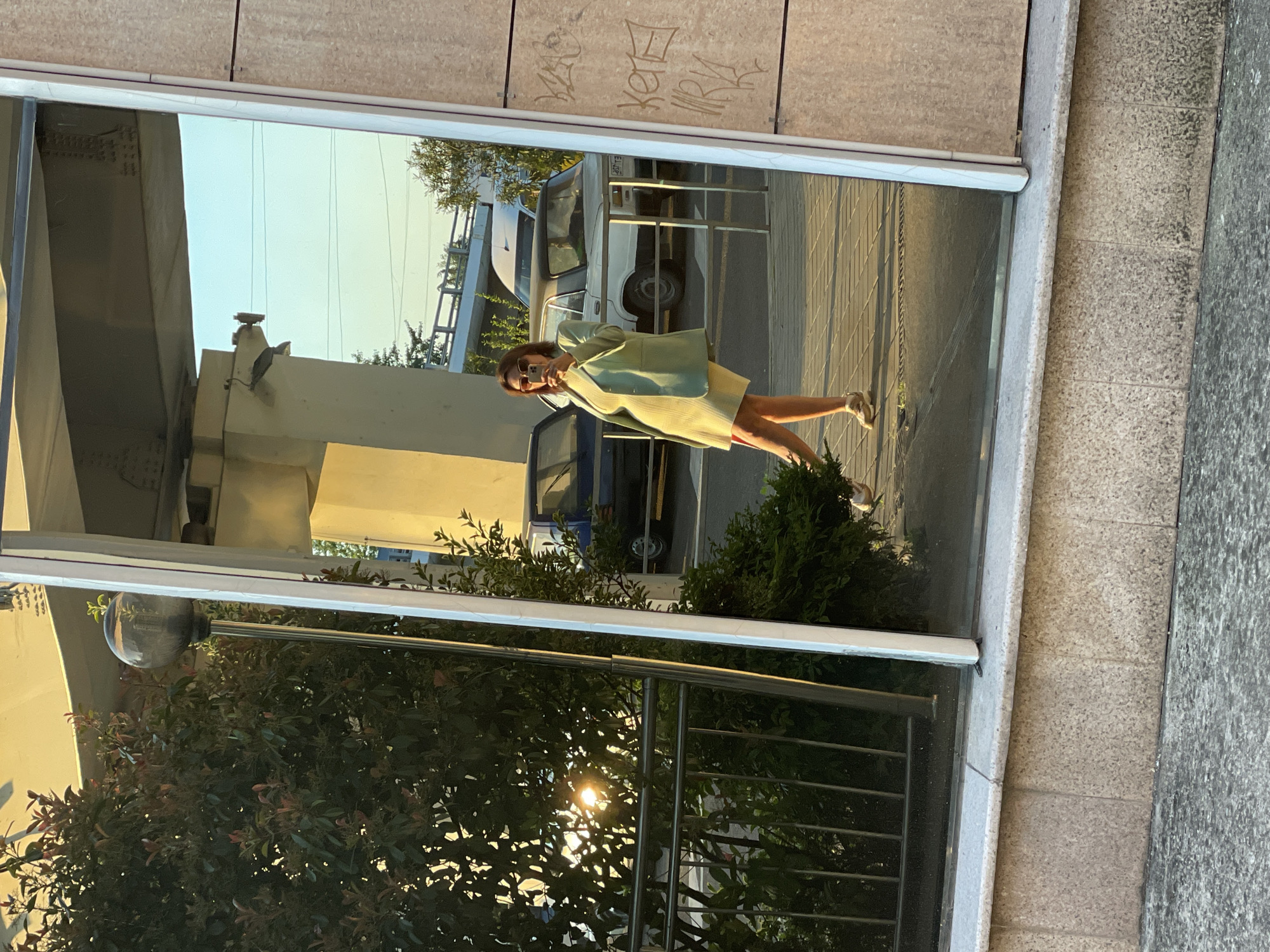 The image captures a reflection of a person taking a selfie in a large window or mirrored surface on a building. The original media file is rotated 90 degrees counter-clockwise, with the person appearing on their side. When viewed in the correct orientation, the reflection shows an outdoor, urban scene.
The primary subject is a young woman, likely in her 20s or 30s, who is seen in the reflection holding a smartphone to take a selfie. She has brown hair and is wearing sunglasses. Her outfit consists of a light-colored (possibly mint green or light yellow-green) long-sleeved top or light jacket and a matching or similarly colored skirt or dress that falls above the knee. She is wearing light-colored athletic shoes. Her posture is casual, with one leg slightly bent.
The reflected background behind the woman includes a street with what appears to be a white van or similar vehicle and part of a blue vehicle. Metal railings or barriers are also visible on the street. The sky, appearing light blue to pale yellow, is reflected above the vehicles. Towards the bottom right of the reflection, bright sunlight pierces through green foliage, creating a noticeable sun flare. Bushes and other greenery are also reflected at the lower edge of the window.
The actual building surrounding the reflective surface features light-colored, possibly granite or concrete, tiled walls. Above the window, on the building's exterior, there is visible graffiti in white and black. One clear tag reads "KOTE" (КОТЭ in Cyrillic, meaning "kitten" or "kitty"). To the right of the window, the ground transitions from light-colored paving stones to a darker, possibly asphalt or textured concrete, surface.
The lighting suggests late afternoon or early evening, often referred to as golden hour, given the warm tones and strong highlights from the sun. The scene takes place in an urban setting, consistent with the provided location context of Sochi, Russia.