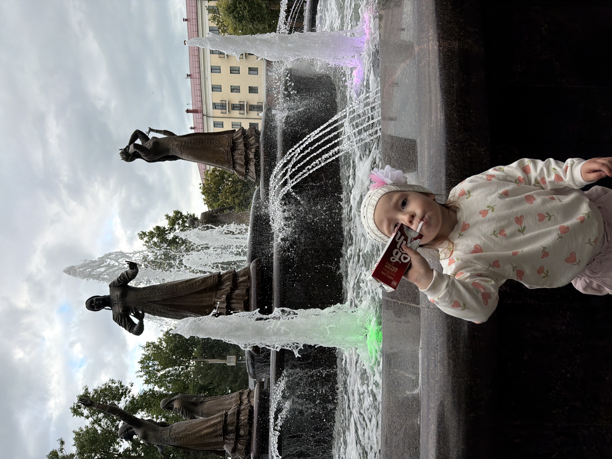 The image shows a young child wearing a white knitted hat with a flower, and a white sweatshirt with a floral pattern. The child is holding a red juice box and appears to be looking at the camera. The child is positioned in front of a fountain with several tiers and water jets. In the background, there are bronze statues of women and a building with several floors and windows. The sky is overcast. The overall scene appears to be outdoors in a public space, likely a park or plaza, given the fountain and statues. The lighting suggests it is daytime. Text visible on the juice box is in Russian and reads "YOGO" and "ГУСТОЙ ДЕСЕРТ" which translates to "THICK DESSERT". The location context is Ufa, Russia.