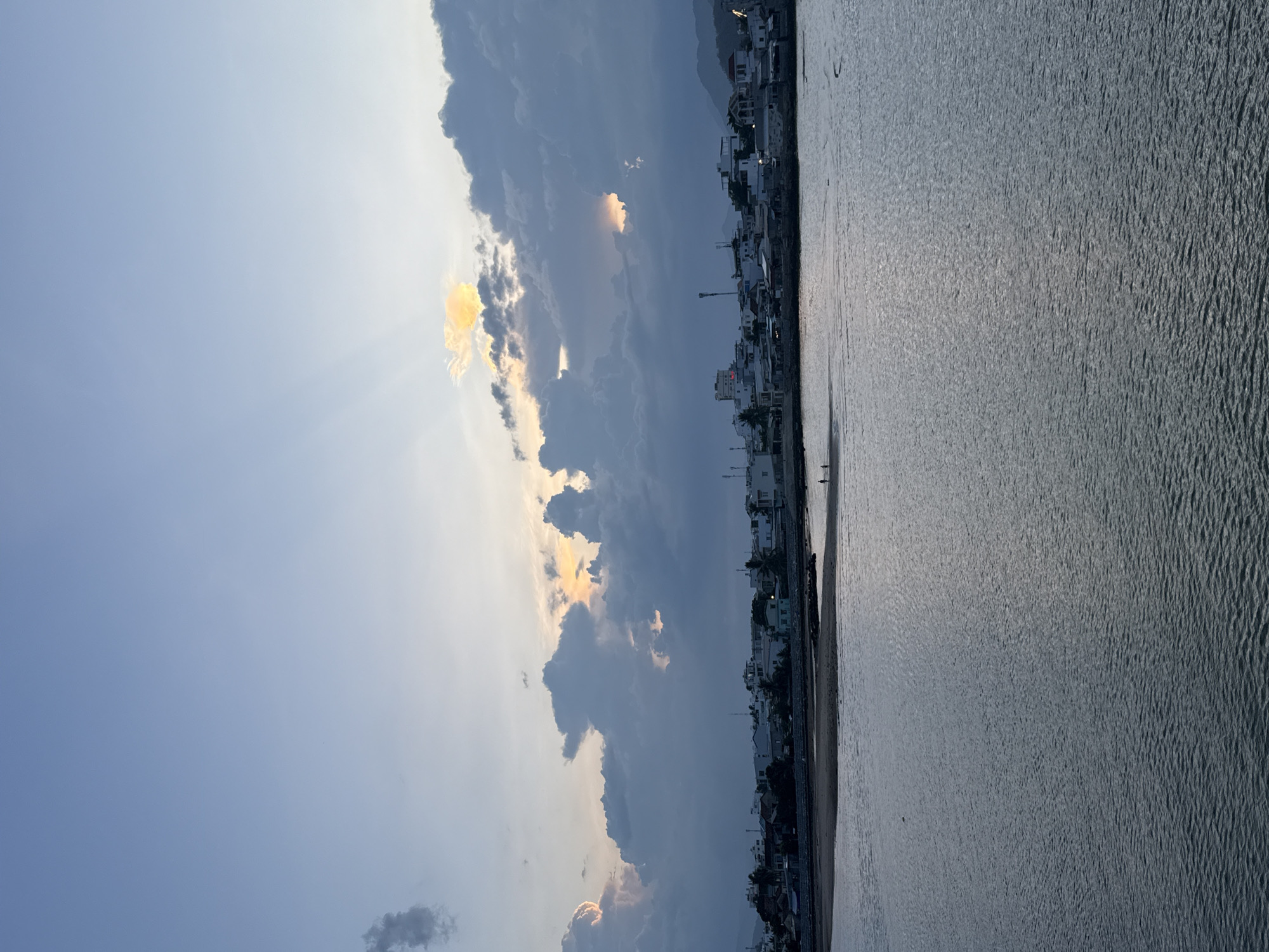 The image captures a serene coastal scene in Nha Trang Ward, Vietnam, likely during the late afternoon or early evening. The foreground is dominated by a calm expanse of water, reflecting the muted tones of the sky and surrounding landscape. Gentle ripples on the water's surface suggest a tranquil environment.
Along the shoreline, a sandy beach is visible, with a few indistinct figures walking, adding a sense of human presence to the scene. Beyond the beach lies a densely packed settlement of buildings, characterized by white and light-colored structures, indicative of urban development along the coast. Some of these buildings appear to be residential or commercial properties. In the background, the silhouette of hills or mountains can be observed, providing a natural backdrop to the urban and coastal elements.
The sky is a significant feature of the image, with dramatic cloud formations and shafts of sunlight breaking through. The clouds are a mix of dark, heavy masses and lighter, more diffused sections. Sunlight emanates from behind the clouds, creating rays that cast a soft glow on the scene, particularly illuminating a patch of sky with a warm, golden hue. The overall color palette is a blend of blues, grays, and subtle warm tones from the sunlight, suggesting a transitional time of day. The atmosphere appears peaceful and somewhat atmospheric due to the interplay of light and clouds.