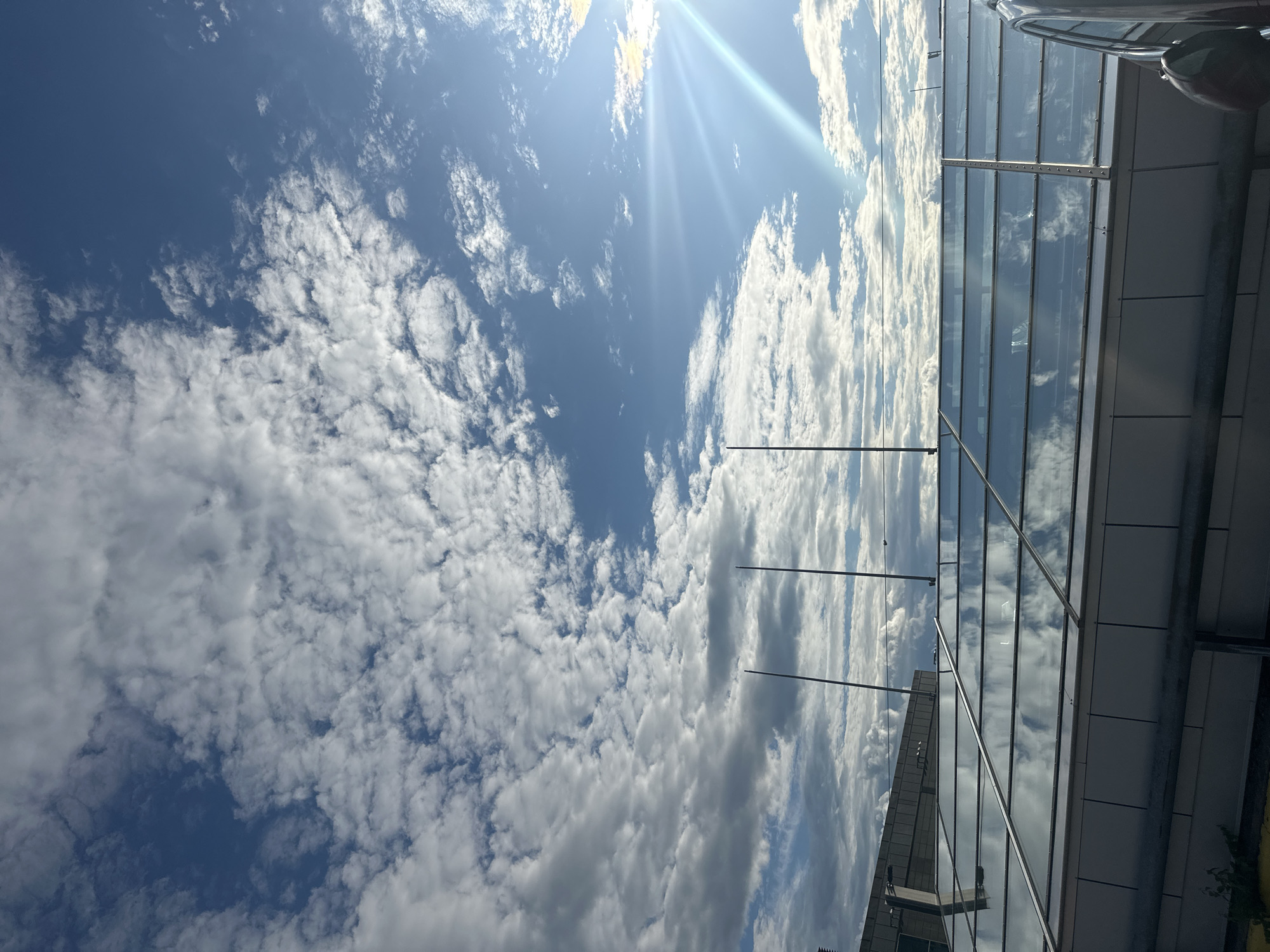 A wide-angle outdoor shot captures a bright, sunlit sky filled with scattered white clouds. The sun is positioned in the upper right corner, casting bright rays and lens flare. Below the sky is a modern building with a glass roof, reflecting the clouds and sky. The roof is supported by metal beams. Three tall, thin poles extend upwards from behind the glass roof. Part of a dark-colored vehicle is visible on the right side of the image. In the lower left, some greenery and a yellow surface are partially visible. The scene appears to be in an urban or commercial area.