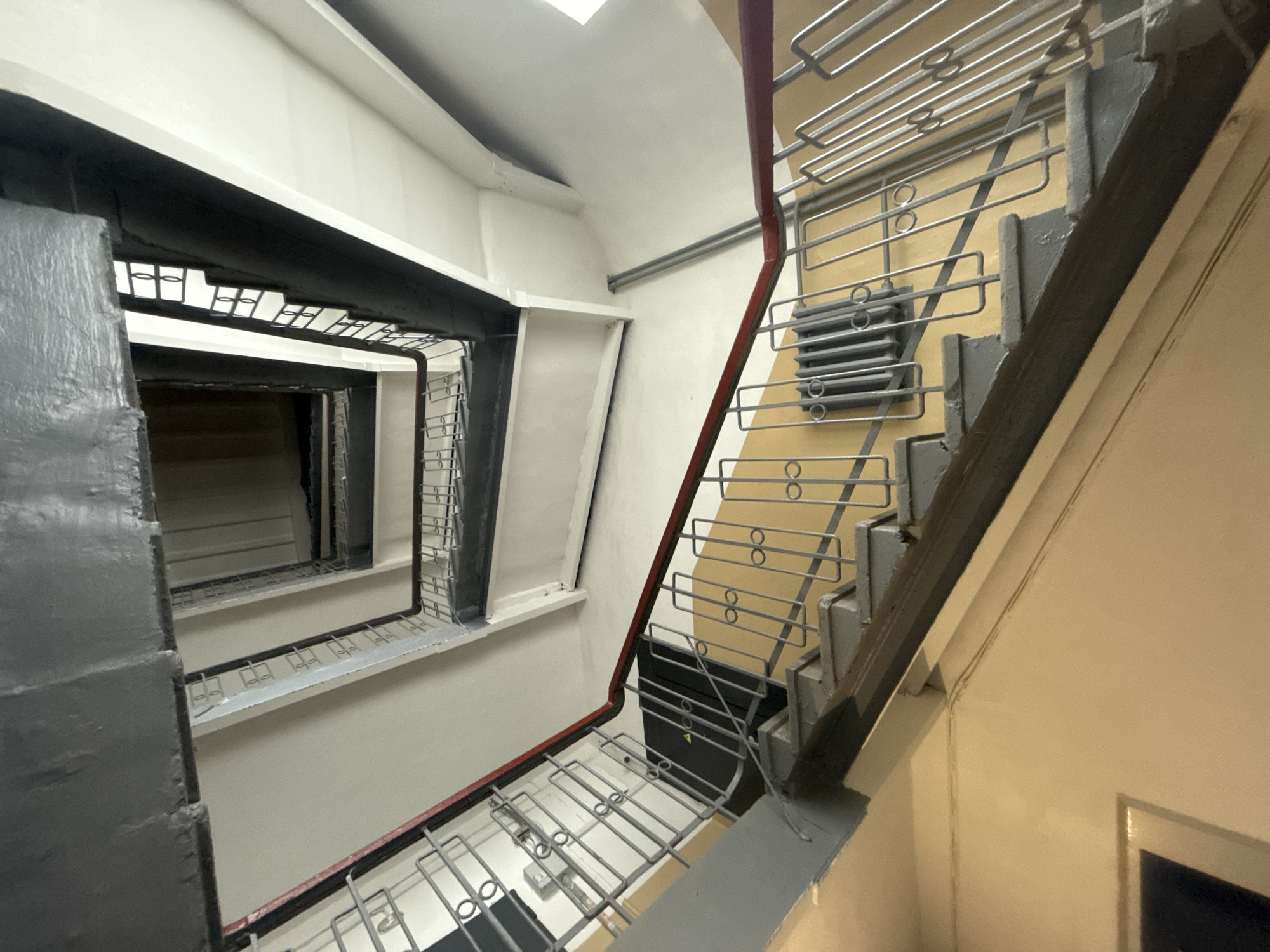The image depicts an interior staircase in Yekaterinburg, Russia. The staircase is constructed with a metal railing system, featuring horizontal bars and circular decorative elements. The walls are painted in neutral tones, with contrasting dark gray or black trim around the stairwell openings. The staircase extends downwards into the building, suggesting multiple floors. The lighting is artificial and comes from an overhead source.