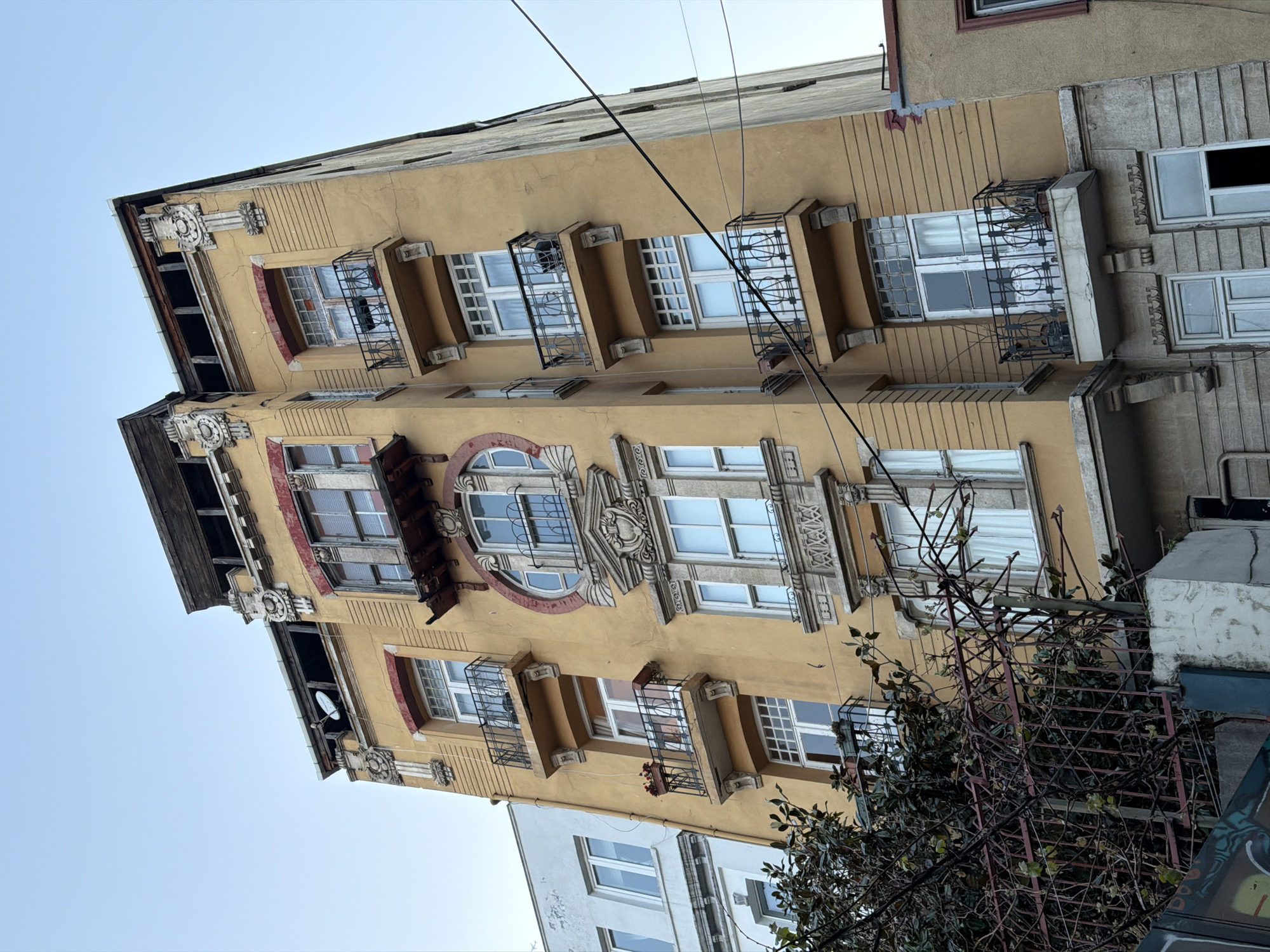 The image displays a tall, narrow building with a light yellow or ochre-colored facade, suggesting an urban setting characteristic of areas like Beyoğlu, Turkey. The building shows signs of age, with visible cracks on its surface and some worn architectural details.
The structure features multiple floors, each adorned with rectangular windows. Many of these windows are accompanied by small, decorative wrought-iron balconies or protective grilles, some extending slightly outward. A particularly ornate section on a lower-middle floor stands out, featuring a large, central arched window framed by elaborate relief carvings, decorative cornices, and engaged columns. The surrounding window frames are often painted in a contrasting reddish-brown color.
Adjacent buildings are partially visible: a simpler white structure with modern windows is seen at the bottom left, and a greyer building with similar window grilles and architectural elements appears to the right. Utility wires or power lines traverse the clear blue sky above the building. On the upper left side of the main building, a small satellite dish is mounted. At the bottom right, dark, tangled branches of a tree or bush are seen behind a metal fence or trellis.
The scene is captured during the daytime under a bright, cloudless sky, indicating fair weather. There is no human activity or visible text within the frame.