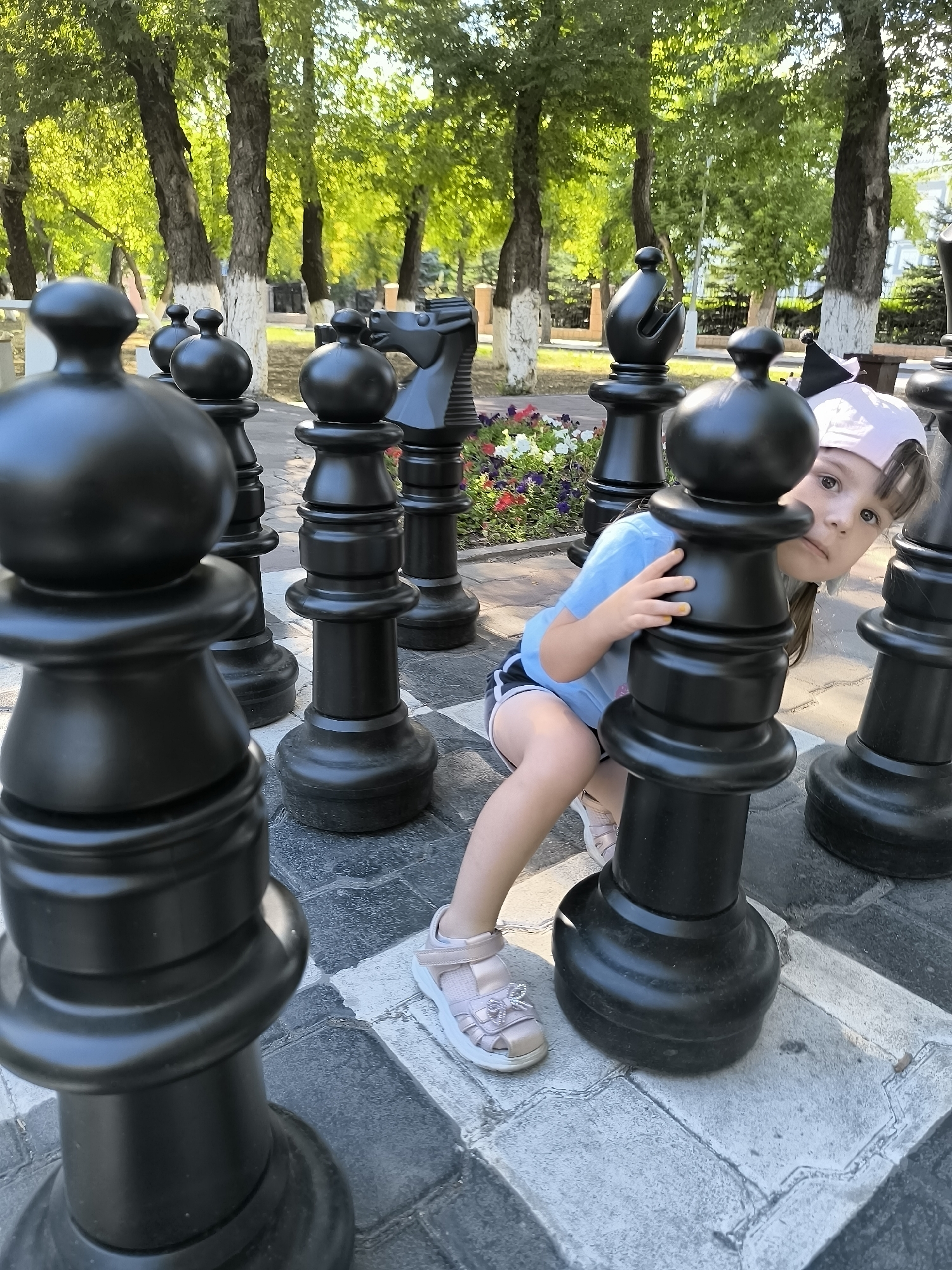 A young girl with a pink baseball cap and blue t-shirt is crouching behind a large black chess pawn in an outdoor park. She is peeking out from behind the pawn with a playful expression. She is wearing light pink sandals. The giant chess pieces are arranged on a checkered ground in what appears to be a city park. In the background, there are trees with green leaves and a flower bed with colorful flowers. The scene is illuminated by natural sunlight. This photograph was taken in Karaganda, Kazakhstan.