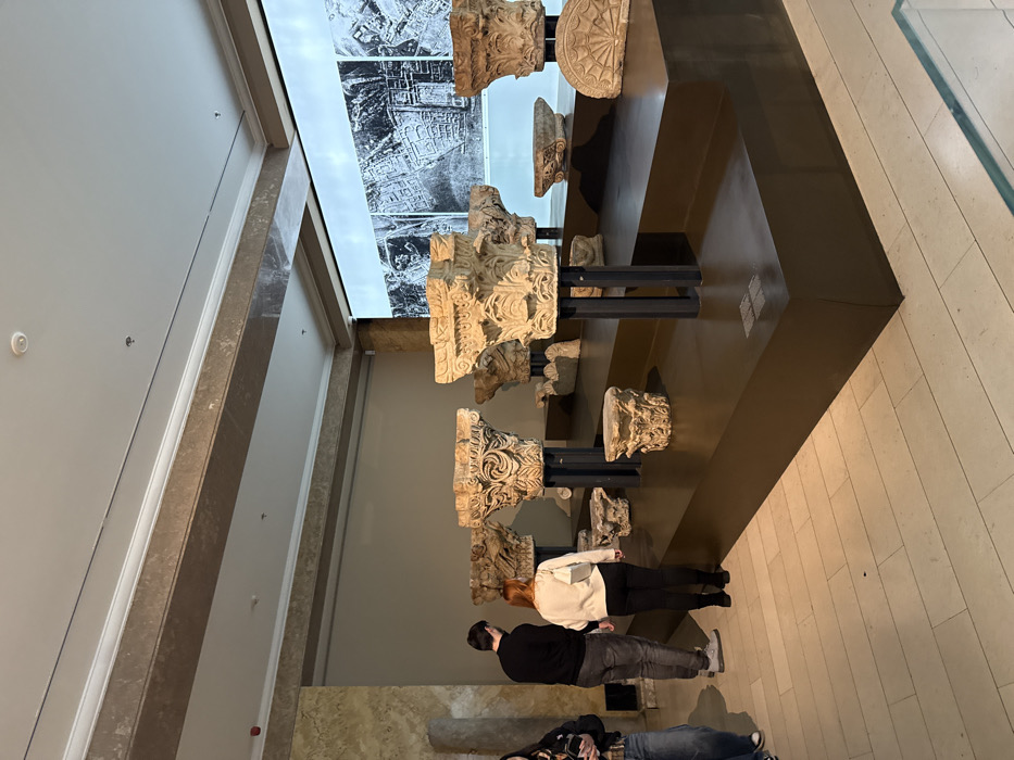 The image captures an interior view of an exhibition hall located within the National Museum of Serbia in Belgrade. The scene presents a spacious room with a high, light-colored ceiling featuring recessed lighting and decorative cornices and beams. A large, multi-tiered dark brown display platform dominates the mid-ground, extending towards the right.
Approximately ten to fifteen ancient stone architectural elements are exhibited on this platform, primarily consisting of ornate column capitals, many displaying carved acanthus leaves characteristic of Corinthian or composite styles. Some capitals are mounted on dark vertical metal supports, while others rest directly on the display surface or smaller risers. Additionally, fragments such as a semi-circular stone relief with radiating lines are visible.
A prominent feature on the rear wall is a large illuminated screen or projection displaying a detailed monochrome aerial photograph or historical map of a city, depicting a grid-like urban layout with visible structures. The left wall features a section of textured, light-toned stone paneling. The floor is covered with light-colored rectangular tiles arranged in a staggered pattern.
Three individuals are present on the left side of the frame. A man, seen from behind, wears a black long-sleeved shirt and grey trousers, observing the displayed artifacts. Adjacent to him, a woman with reddish-brown hair, also seen from behind, wears a light-colored sweater and dark trousers, carrying a light shoulder bag, and is similarly engaged in viewing the exhibits. Partially visible on the far left, another individual, dressed in denim jeans and a dark jacket, holds a smartphone. All individuals are passively observing the museum artifacts.