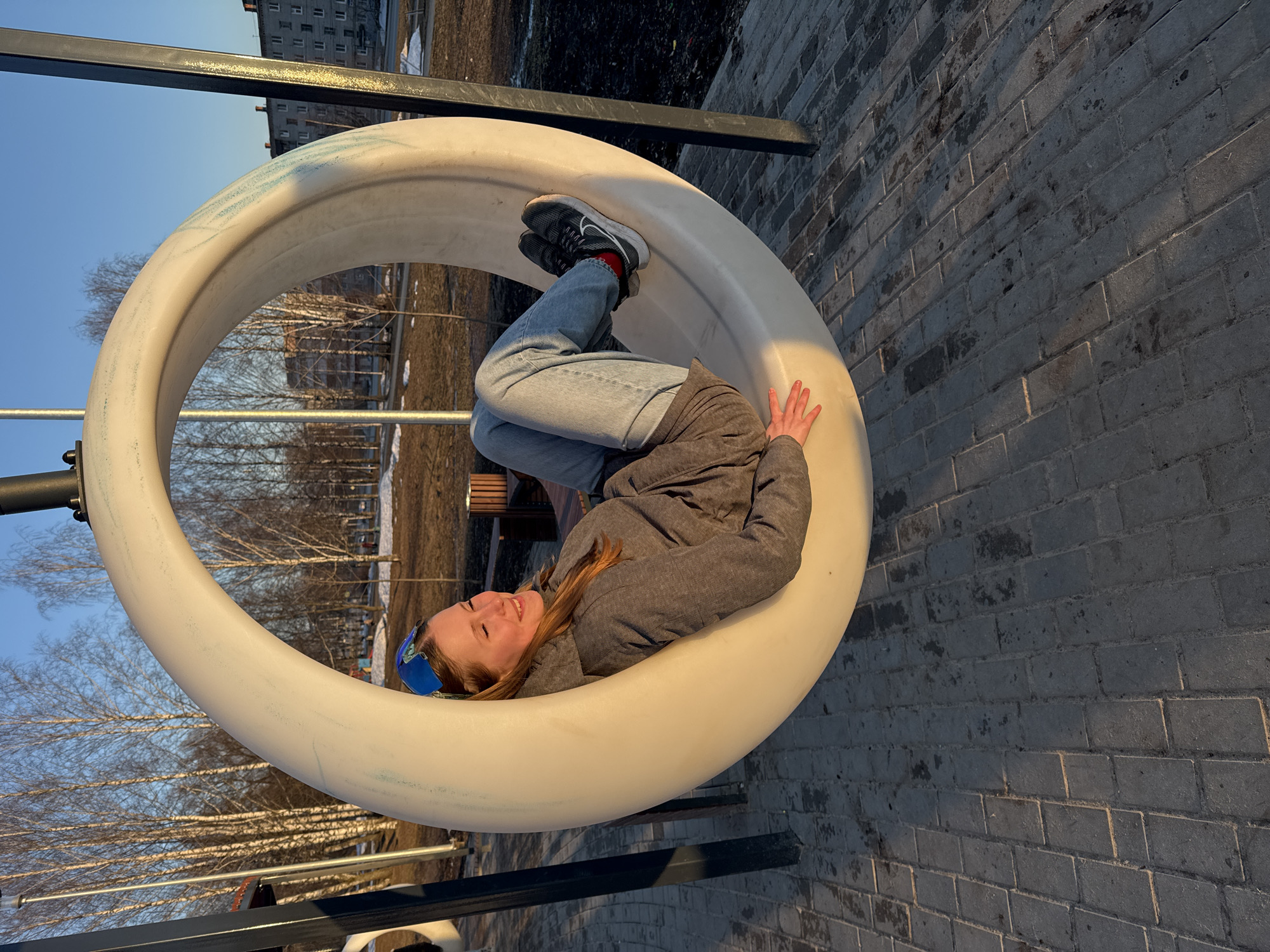 A fair-skinned woman is inside of a large, cylindrical playground apparatus. She is wearing a grey jacket, blue jeans, and athletic shoes with a visible red sock. Her head is tilted to the left, and she has a blue headband. The cylindrical apparatus is white and supported by metal bars. The ground is paved with grey bricks. In the background are bare trees, a building, and patches of snow. The photo appears to have been taken in the city of Berezniki, Russia, based on the style of the buildings and the appearance of the landscape. The lighting suggests it is late afternoon or early morning.