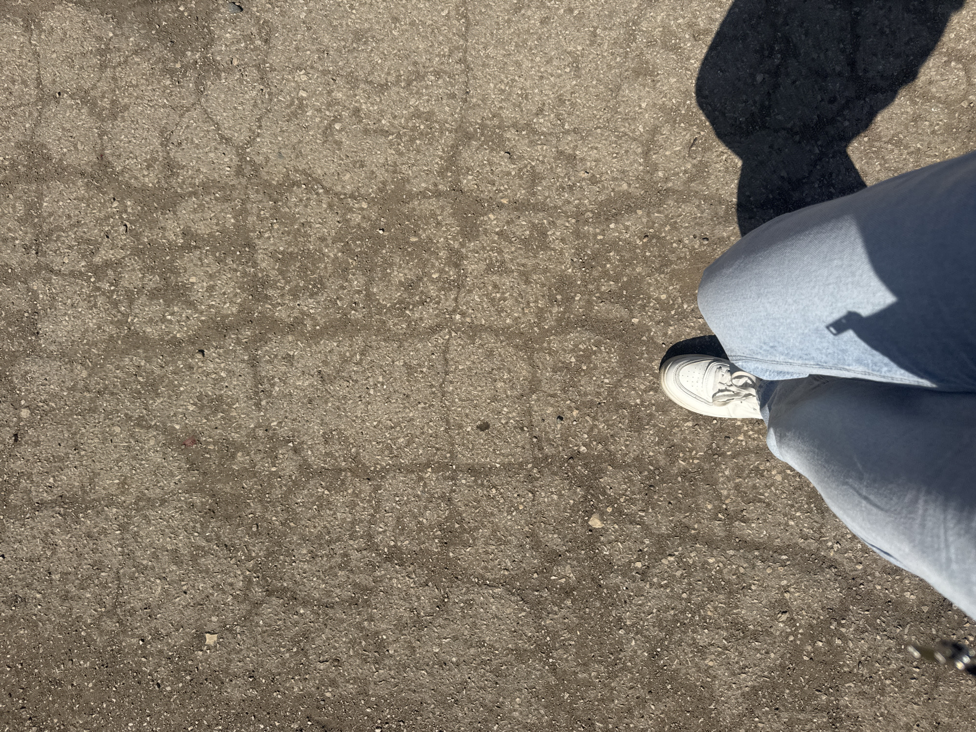 The scene is an asphalt surface, likely a street or pavement. The surface shows wear with cracks and variations in texture. In the upper right quadrant of the frame, the legs of a person are visible, clad in light-blue denim jeans. A single white sneaker is visible on the left foot. A dark, amorphous shadow, presumably cast by the person, is present above the legs. The setting is outdoors in daylight with no other people or objects visible. The location is not determinable from visual information.