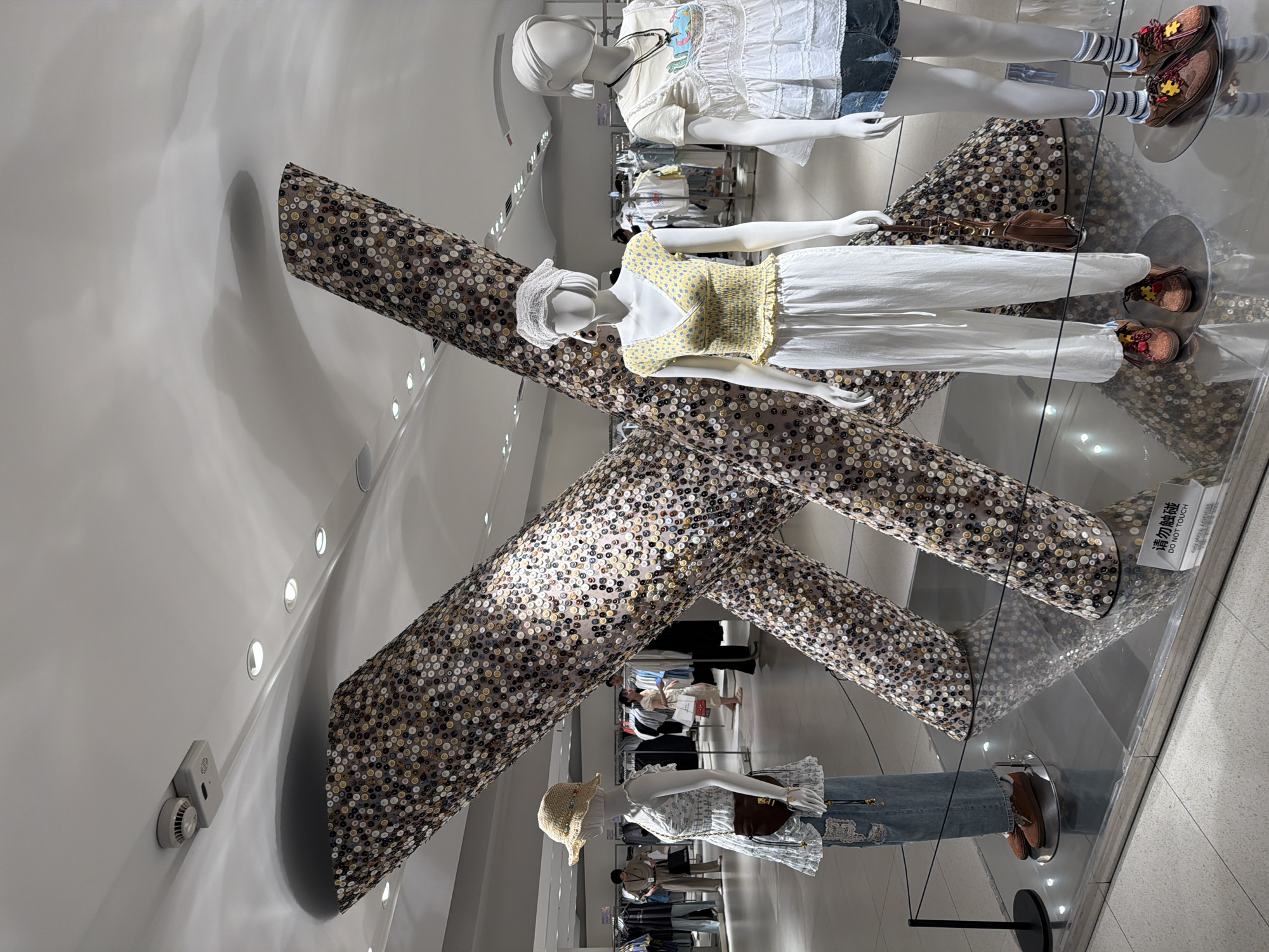 The image shows a retail store interior in Qinhuai District, China, featuring a large "X" shaped art installation made of numerous circular objects, possibly buttons or coins, covering its surface. In front of the installation, three mannequins are displayed. The leftmost mannequin is wearing a light blue floral print dress, a straw hat, and denim pants, with a brown crossbody bag. The middle mannequin is dressed in a yellow floral top with smocking and white wide-leg pants, accessorized with a white headscarf and a brown handbag. The rightmost mannequin wears a white ruffled top with a layered design and denim shorts, with striped socks and brown lace-up shoes. The floor is tiled, and the mannequins are placed on reflective silver circular platforms. In the background, clothing racks and other shoppers are visible, indicating this is a commercial setting. A sign in Chinese and English reading "DO NOT TOUCH" is placed near the base of the art installation.