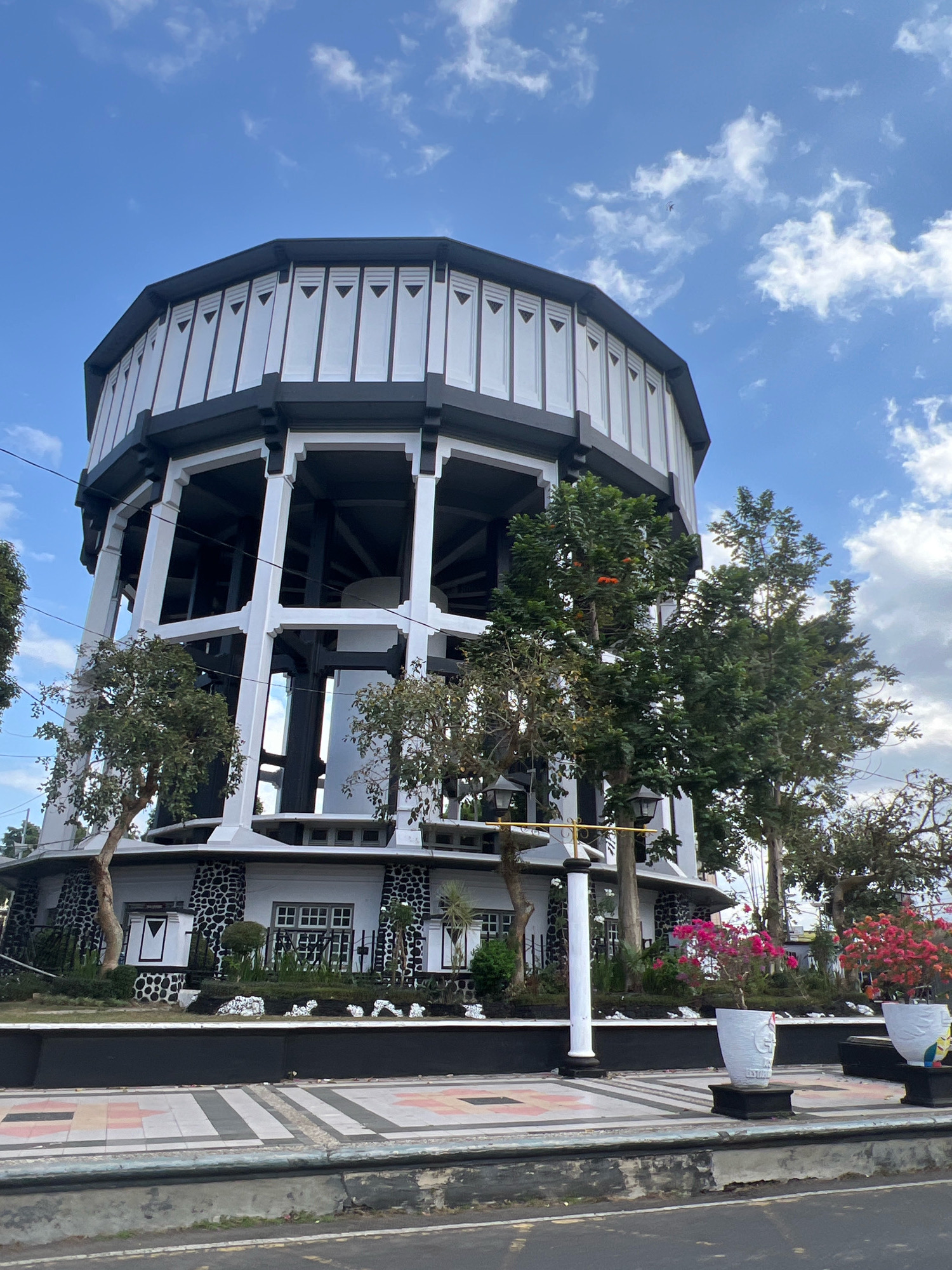 The image shows a tall, octagonal water tower structure in Magelang, Indonesia, with a black roof and white supporting columns. The lower part of the tower is white with black decorative stone patterns and windows. Surrounding the base of the tower are trees, bushes, and flower pots containing pink and red flowers. The foreground features a walkway with decorative patterned tiles, a concrete curb, and a road. The sky is blue with scattered white clouds. The water tower is a prominent landmark, likely situated in a public area or park within the city.