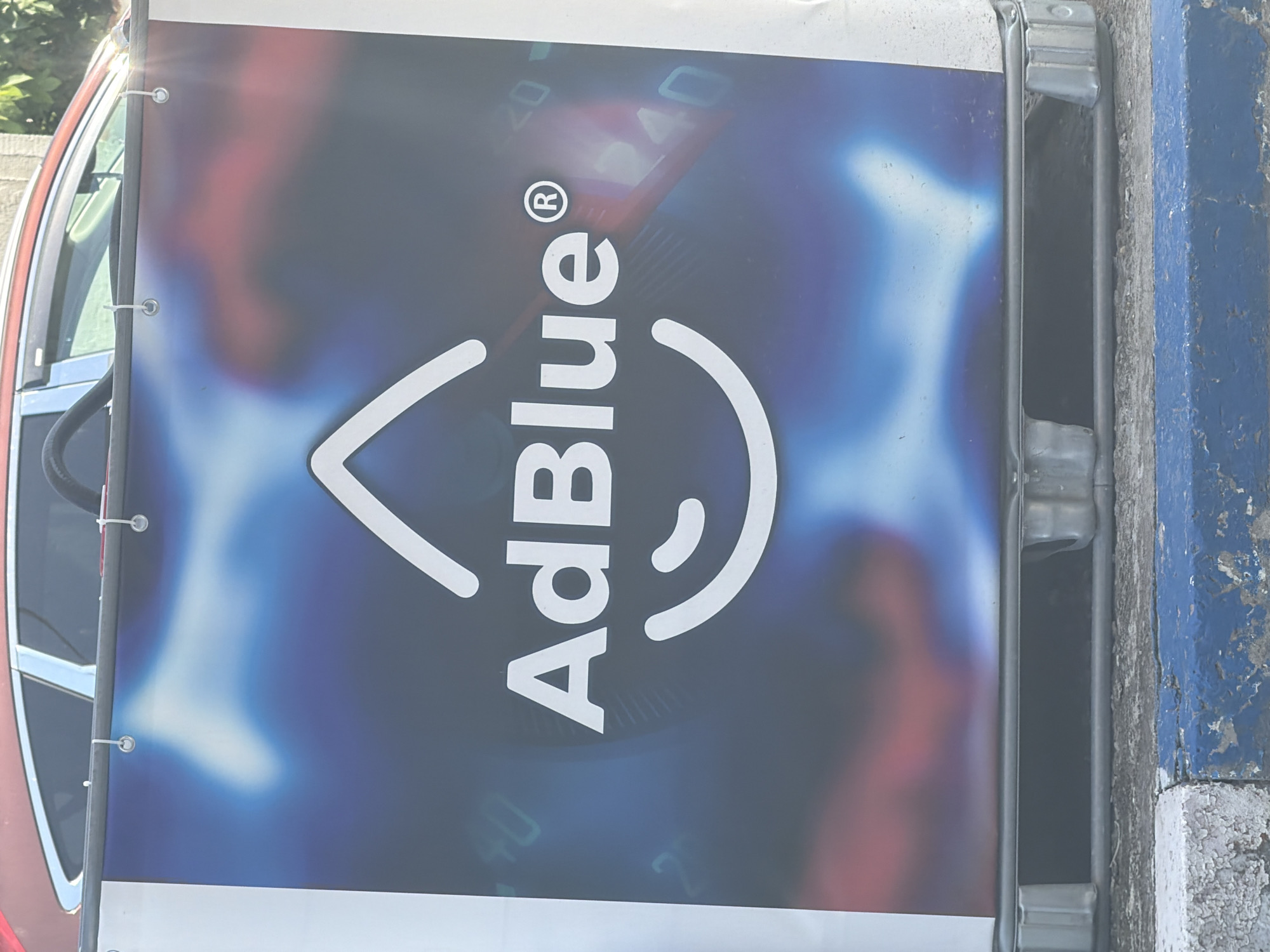 This vertical image shows an outdoor scene featuring a large banner prominently displaying the "AdBlue®" logo and text. The banner, predominantly blue and white, features the word "AdBlue®" in bold white letters, with a stylized white drop symbol above the 'A'. In the blurred background of the banner, faint outlines of numbers, such as "20" and "40", are visible.
The banner is secured along its left edge with visible grommets and white zip ties to a dark vertical pole or frame. Partially visible to the left of the banner is a red car, showing its side window and a chrome trim piece. On the right side of the image, a worn concrete structure, possibly a curb or part of a building foundation, is present. This structure is painted in faded blue and grey, with some areas of chipped paint.
The scene appears to be set during the daytime under bright lighting conditions. No people are visible, and no specific activity is taking place. The overall impression is a static advertisement in an outdoor setting, likely near a road or service area in Shumen, Bulgaria.