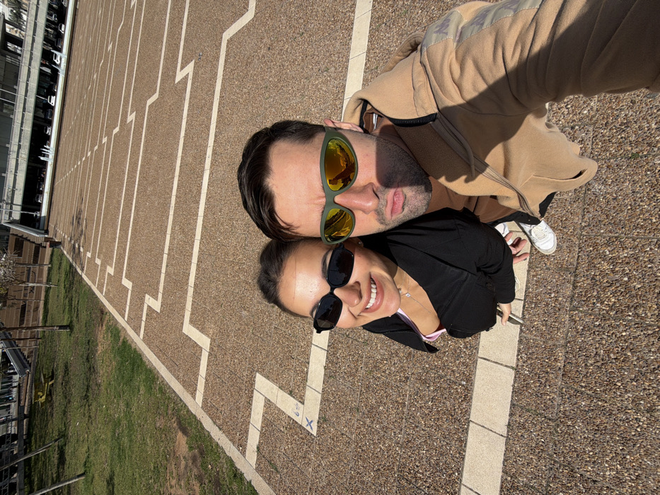 This image captures two individuals, a female and a male, posing for a selfie outdoors in Rabin Square (Kikar Rabin) in Tel-Aviv, Israel.
The female, positioned on the left, has dark hair and wears black sunglasses, a black long-sleeved top over a pink garment, and a silver necklace. She is smiling broadly. The male, on the right, has dark hair and wears green-framed mirrored sunglasses, a tan zippered hoodie over a brown shirt, and has his lips in a puckering expression. Both subjects are looking upward, suggesting the male is holding the camera.
The ground beneath them is a light brown pebbled surface featuring distinctive light-colored geometric patterns, which are characteristic of Rabin Square. To the left of the subjects, a patch of green grass with several small trees and a yellow picnic table is visible. In the background, the multi-level concrete and stone structure of the Tel Aviv City Hall building is prominent, complete with staircases and an upper-level plaza where several vehicles are faintly visible. The scene is illuminated by bright daylight, casting shadows on the subjects and ground.