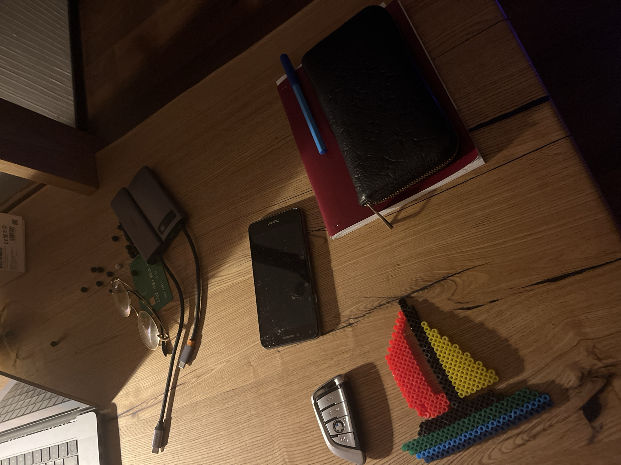 The image depicts a wooden desk surface with various objects, suggesting a workspace or personal area. In the upper left, part of a silver laptop is visible. Several USB cables and adapters are connected to a charging hub or dock. A pair of eyeglasses with gold frames rests on the desk, near a green credit card. A few small, dark, round objects, possibly beads or berries, are scattered around this area. To the right of the glasses, two dark gray power banks are stacked.
In the center of the image, a black smartphone with a cracked screen lies flat. To its left is a silver car key fob with the BMW logo. Below the smartphone, a colorful mosaic pattern made of small beads in blue, green, red, black, and yellow is arranged on the desk.
In the lower right section of the image, a black leather wallet with a textured pattern is placed on top of a red notebook. A blue pen rests on the notebook, positioned above the wallet.
The lighting in the image suggests an indoor setting, possibly at dusk or with artificial lighting, casting shadows on the wooden surface. The overall impression is one of a collection of personal electronic devices, accessories, and craft materials. There are no people visible in the image. The location is described as Ореховка, Russia, which is an urban locality.