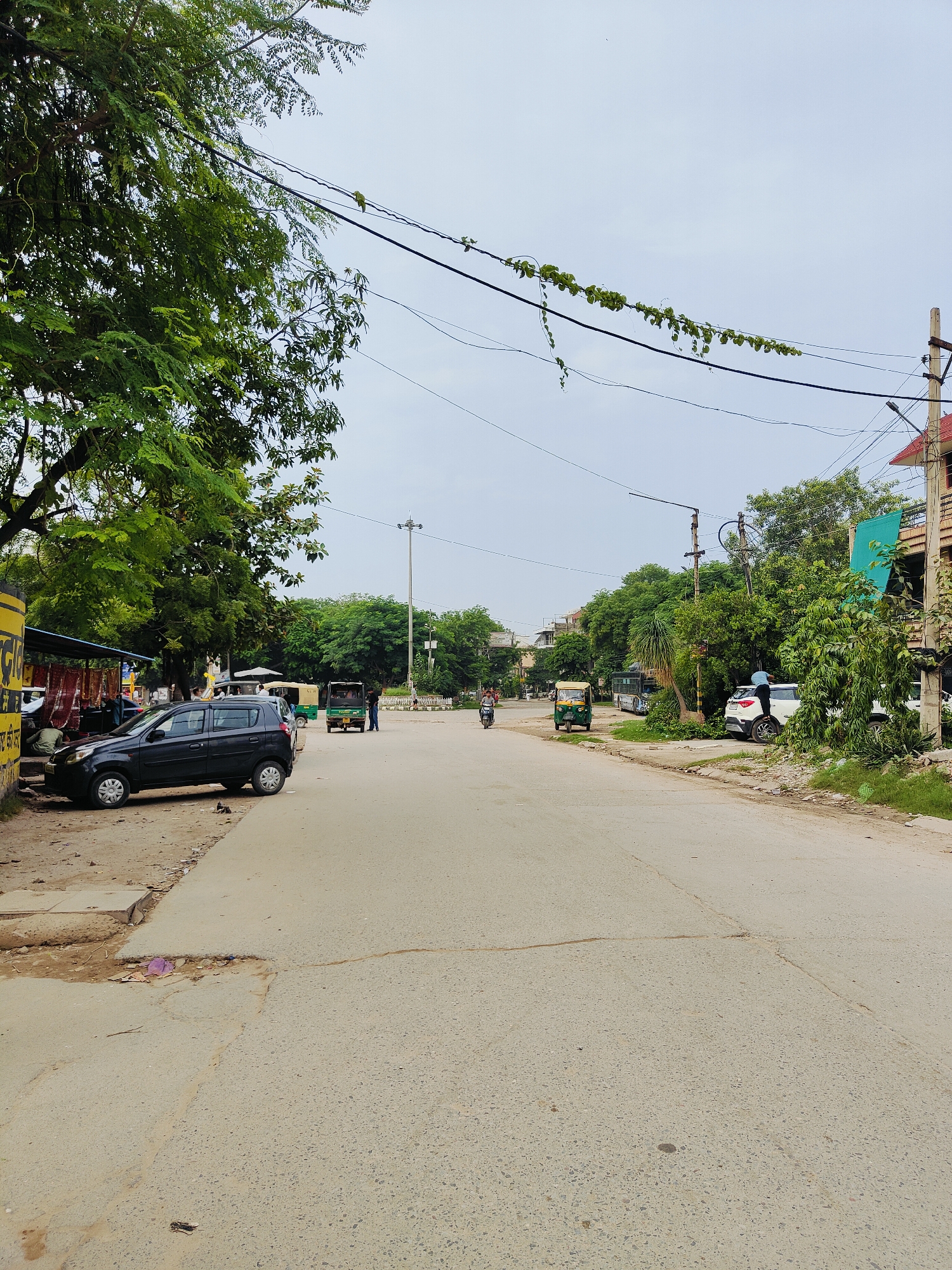 The image depicts a street scene in Gurgaon, India, on an overcast day. The foreground shows a paved road with some cracks and patches, leading into the distance. On the left side of the road, a black car is parked next to a yellow-painted structure with a blue awning. Several people are visible near this structure, with one appearing to be working on the car. Further down the road, several auto-rickshaws and a bus are present, along with more parked vehicles and trees lining the street. Power lines are strung across the sky, with some vegetation growing on them in places. The overall environment suggests a moderately busy urban street with a mix of commercial and residential elements.