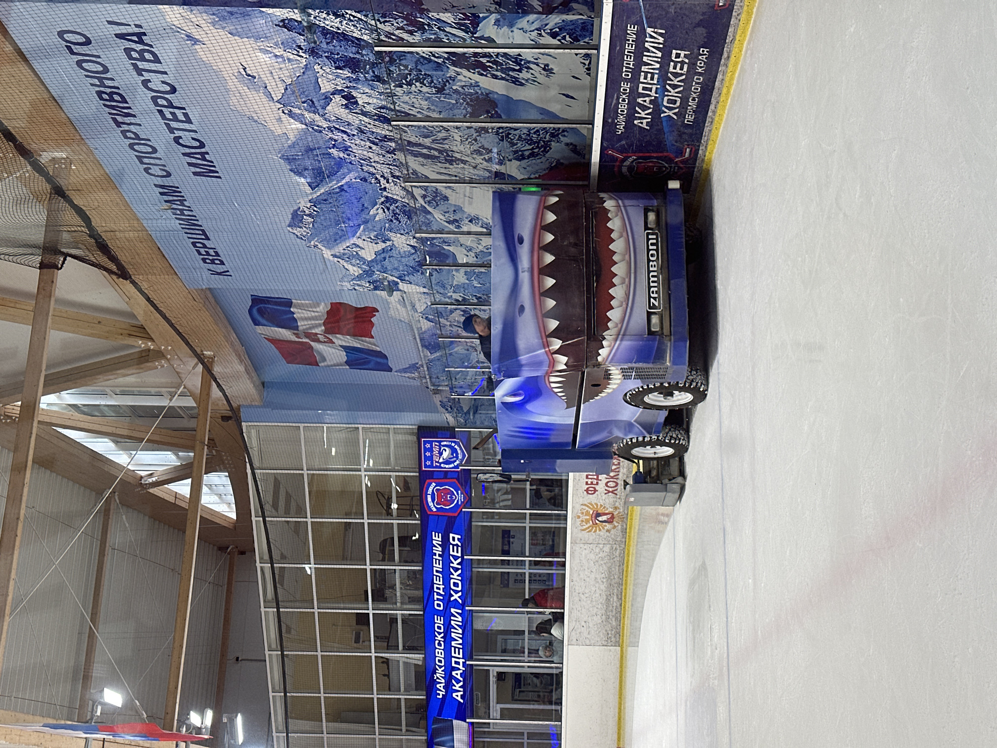 The image depicts the interior of an ice rink in Chaikovsky, Russia, featuring a Zamboni ice resurfacing machine with a distinctive shark-themed design. The Zamboni is positioned on the ice surface, ready for operation. Overlooking the rink are banners and signage in Russian, including "Чайковское Отделение Академии Хоккея" (Chaikovsky Branch of the Hockey Academy), indicating the location of a hockey training facility. A large banner displays a mountain range backdrop and the slogan "К Вершинам Спортивного Мастерства!" (To the Heights of Sportsmanship!). The rink's structure includes a high ceiling with exposed wooden beams and netting. Windows provide natural light. People can be seen through the windows.