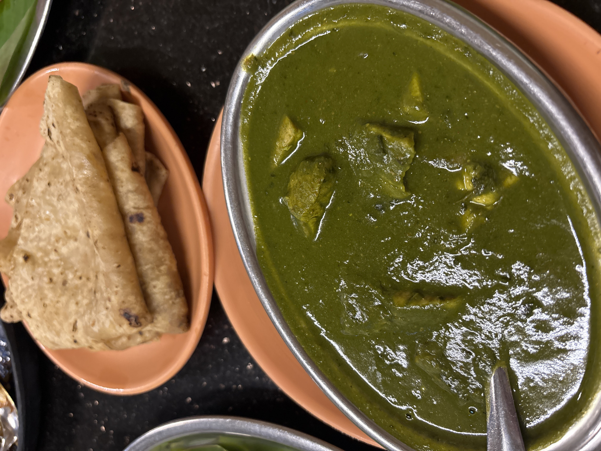 The image shows two dishes of food. On the right, there is a metal bowl filled with a green, thick curry, likely Palak Paneer, an Indian dish made from spinach and cheese. A spoon rests in the curry. To the left, there is an oval orange plate with several rolled flatbreads, possibly roti or chapati. The background is a dark, speckled surface. The scene is likely a restaurant or home meal setting in Kamalapura, India, based on the style of the cuisine and the cuisine type.