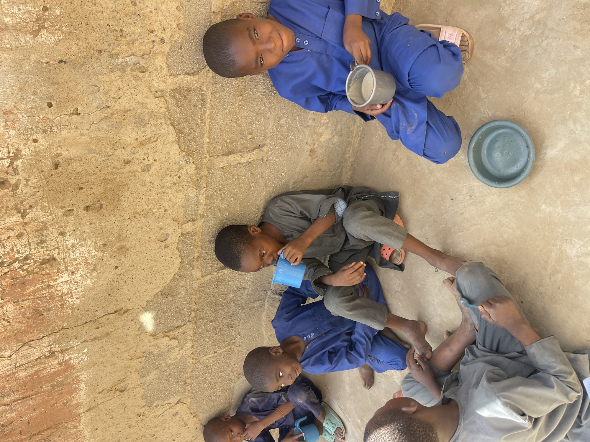 Five children are seated on a concrete surface in what appears to be Unguwar Gabas, Nigeria, judging by the local clothing and housing visible in the background. The surface is light brown, cracked and worn. One child, wearing a blue uniform, sits at the top of the frame holding a metal mug. Beside them is a blue-green bowl. A second child wears gray clothing and holds a blue cup. A third child wears a blue uniform and sits beneath the child in gray. Two other children are at the bottom of the frame: one in blue and one in gray. The children appear to be drinking or preparing to drink from their cups.